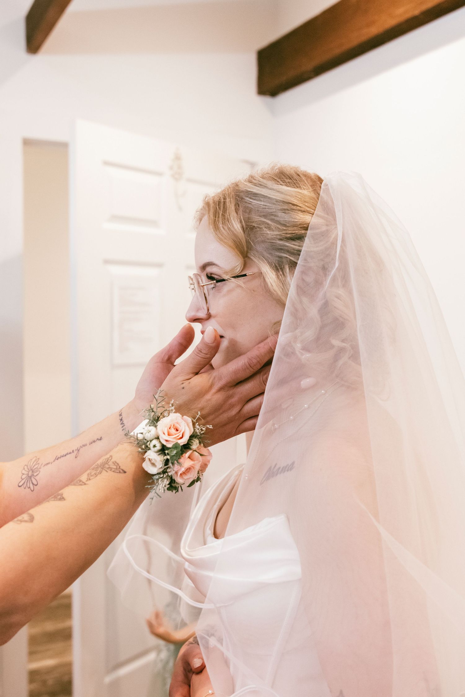 Emotional bride in veil being comforted by tattooed hand wearing pink floral wrist corsage.