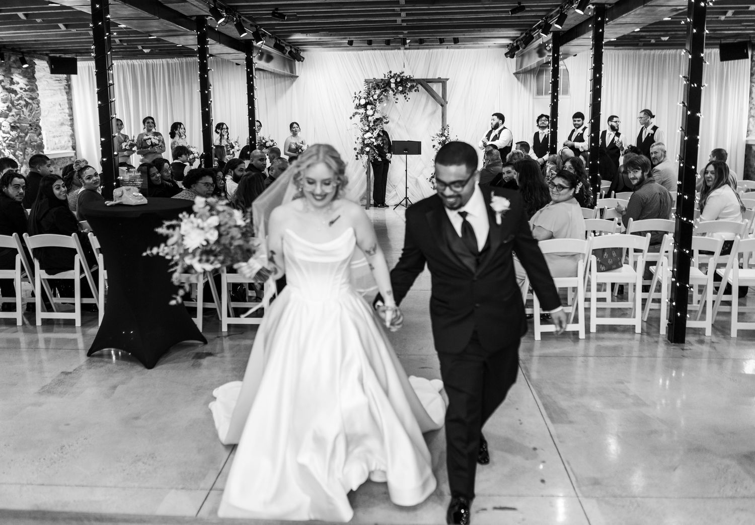 Black and white photo of newlyweds joyfully walking back down the aisle after their indoor wedding ceremony.