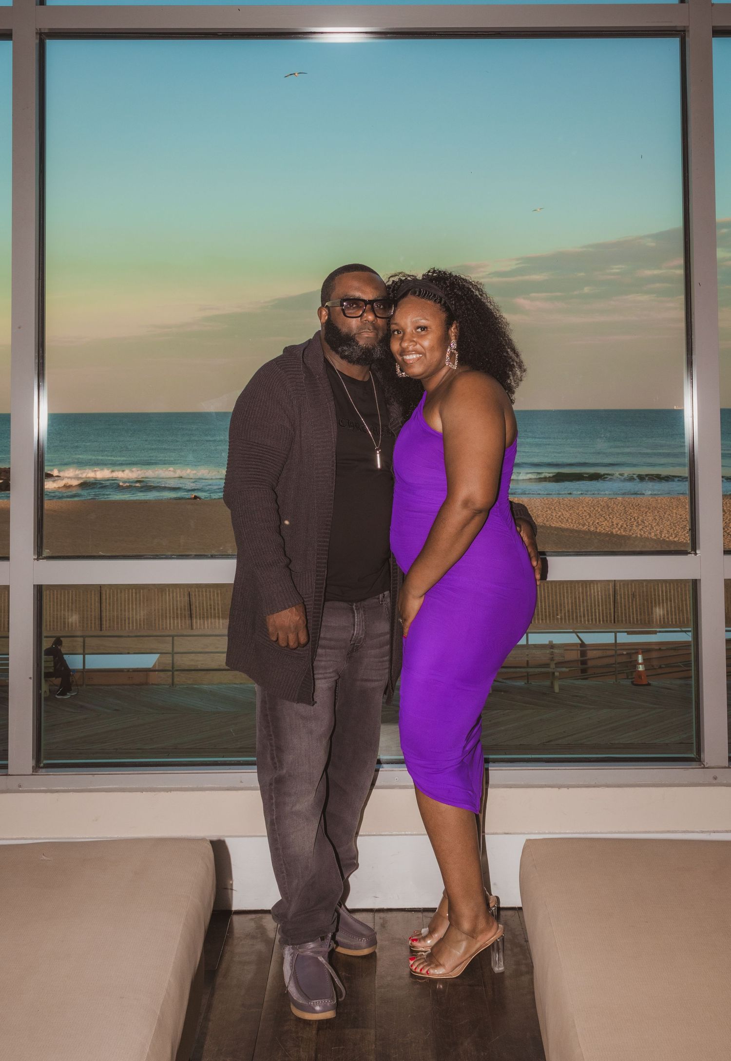 Couple in matching purple dress and gray suit share intimate moments by floor-to-ceiling windows overlooking the ocean at sunset.
