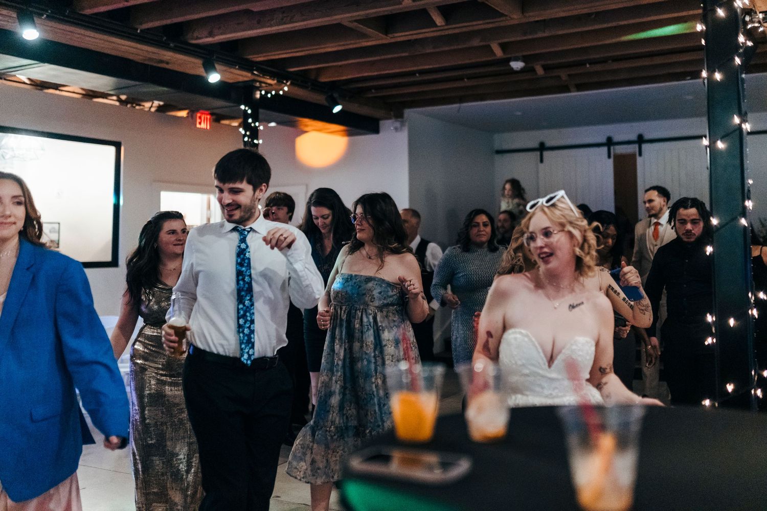 Wedding guests dancing and celebrating at a lively indoor reception with string lights and rustic decor.