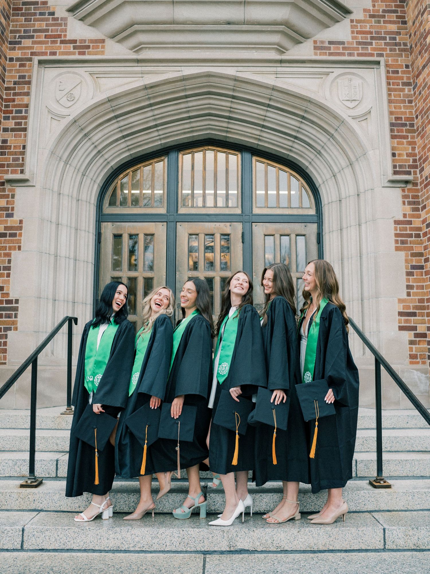 Six female graduates in black and green caps and gowns laughing together on steps outside a stone university building.