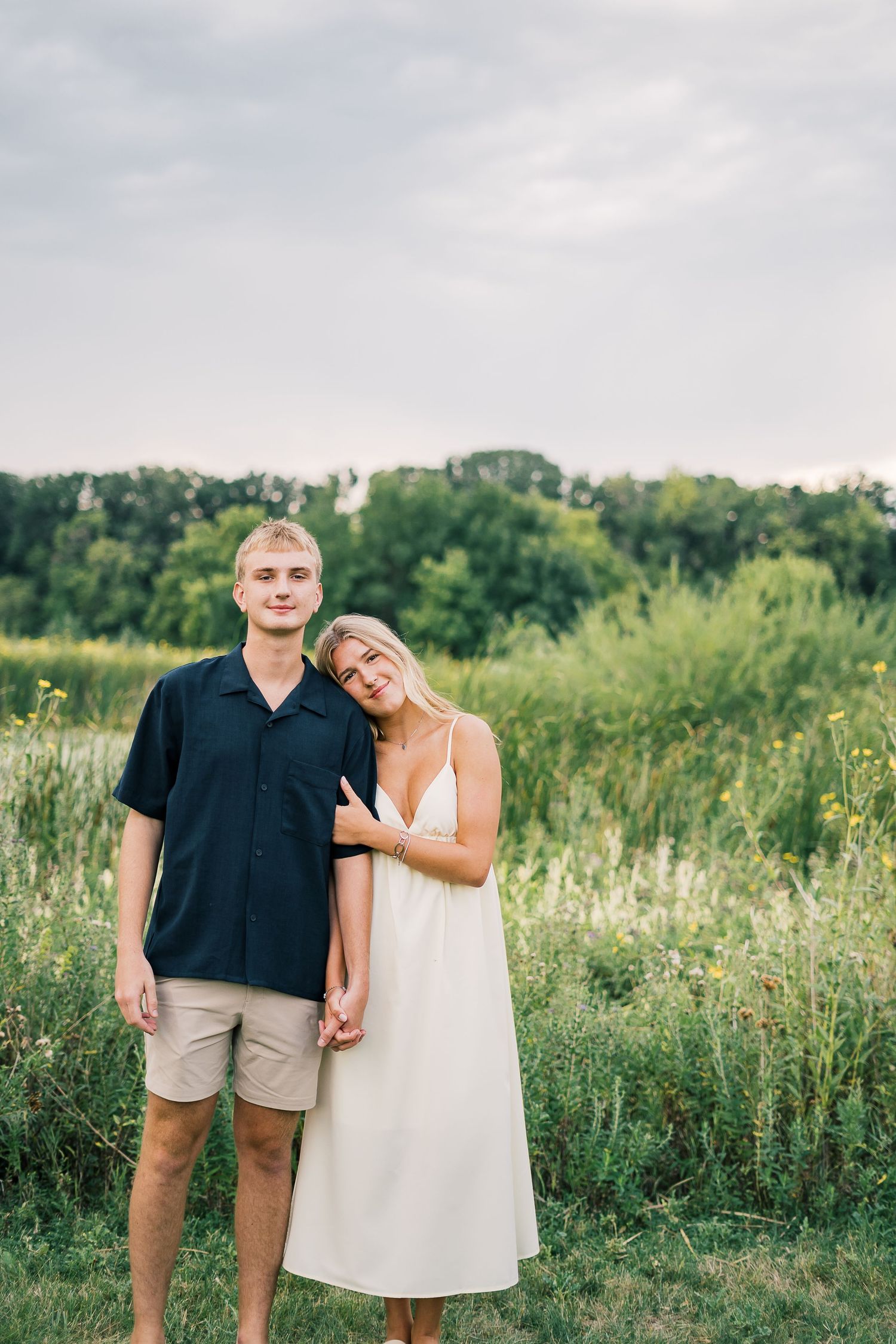 A couple stands together in a sunlit meadow wearing casual summer attire.
