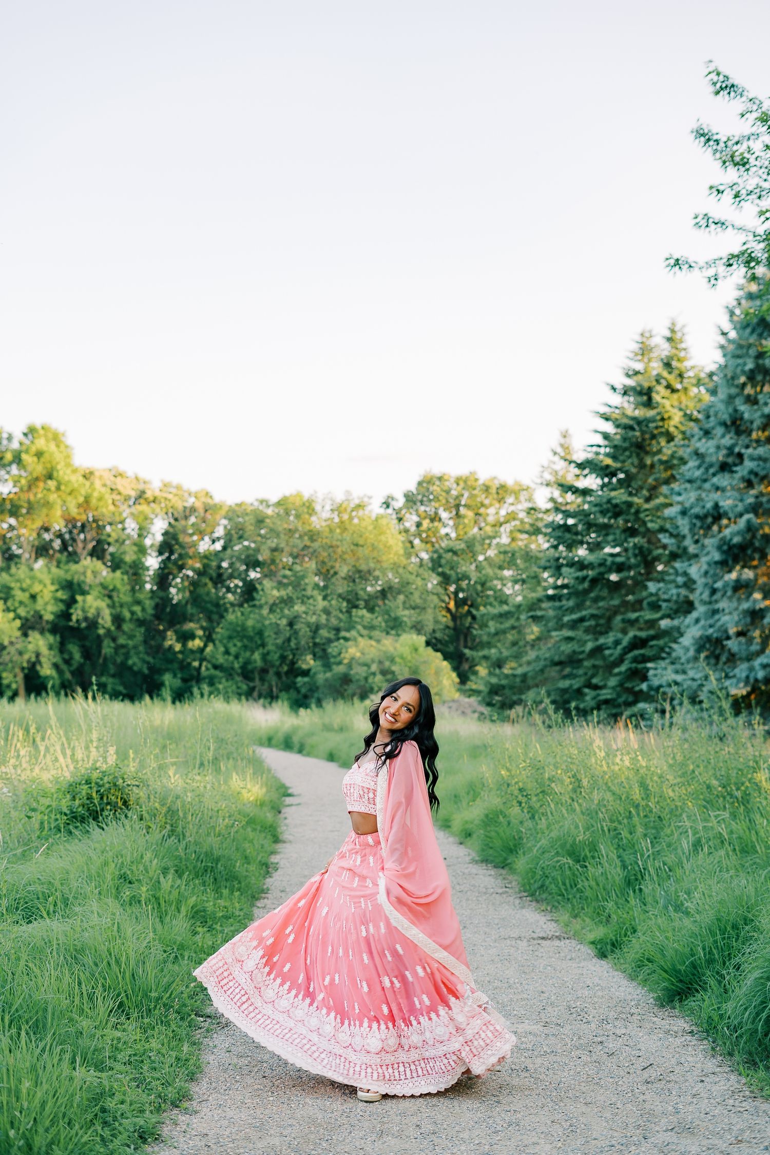 A person in a flowing pink dress twirls joyfully on a nature path surrounded by lush greenery and evergreen trees.