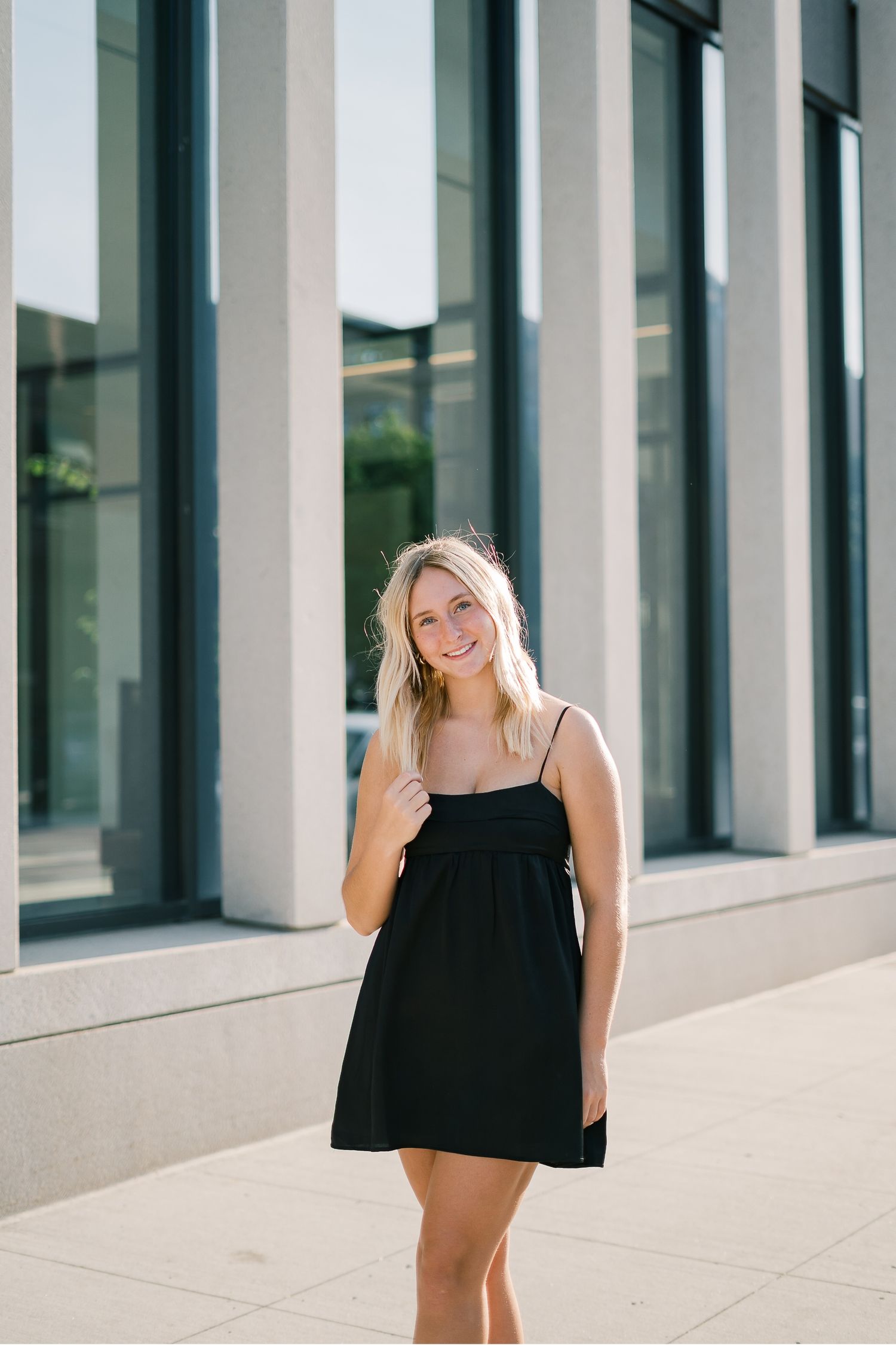 Woman in black dress stands smiling against modern building exterior with white columns and glass windows.
