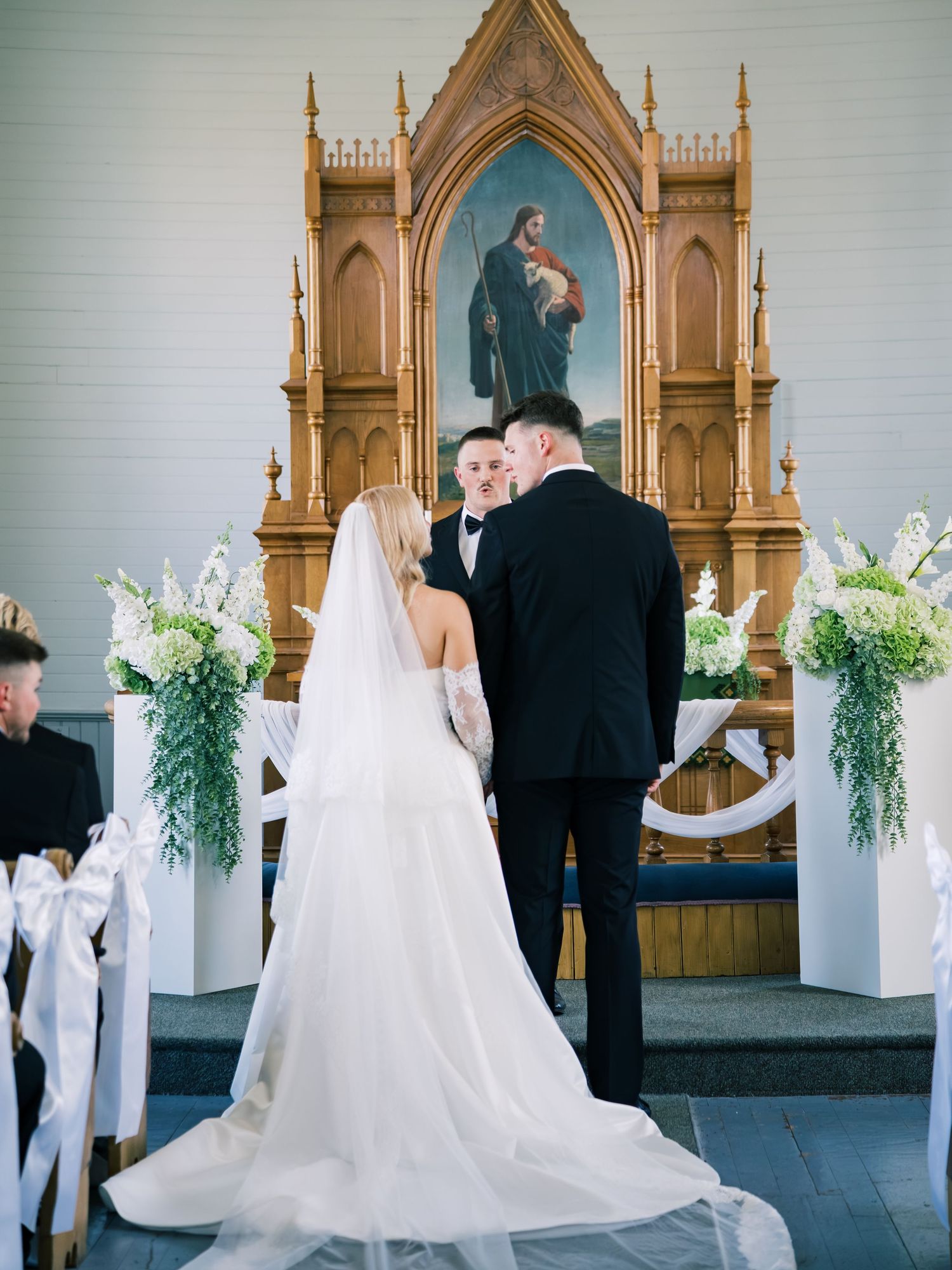 Bride and groom exchange vows at church altar adorned with white floral arrangements and religious painting.