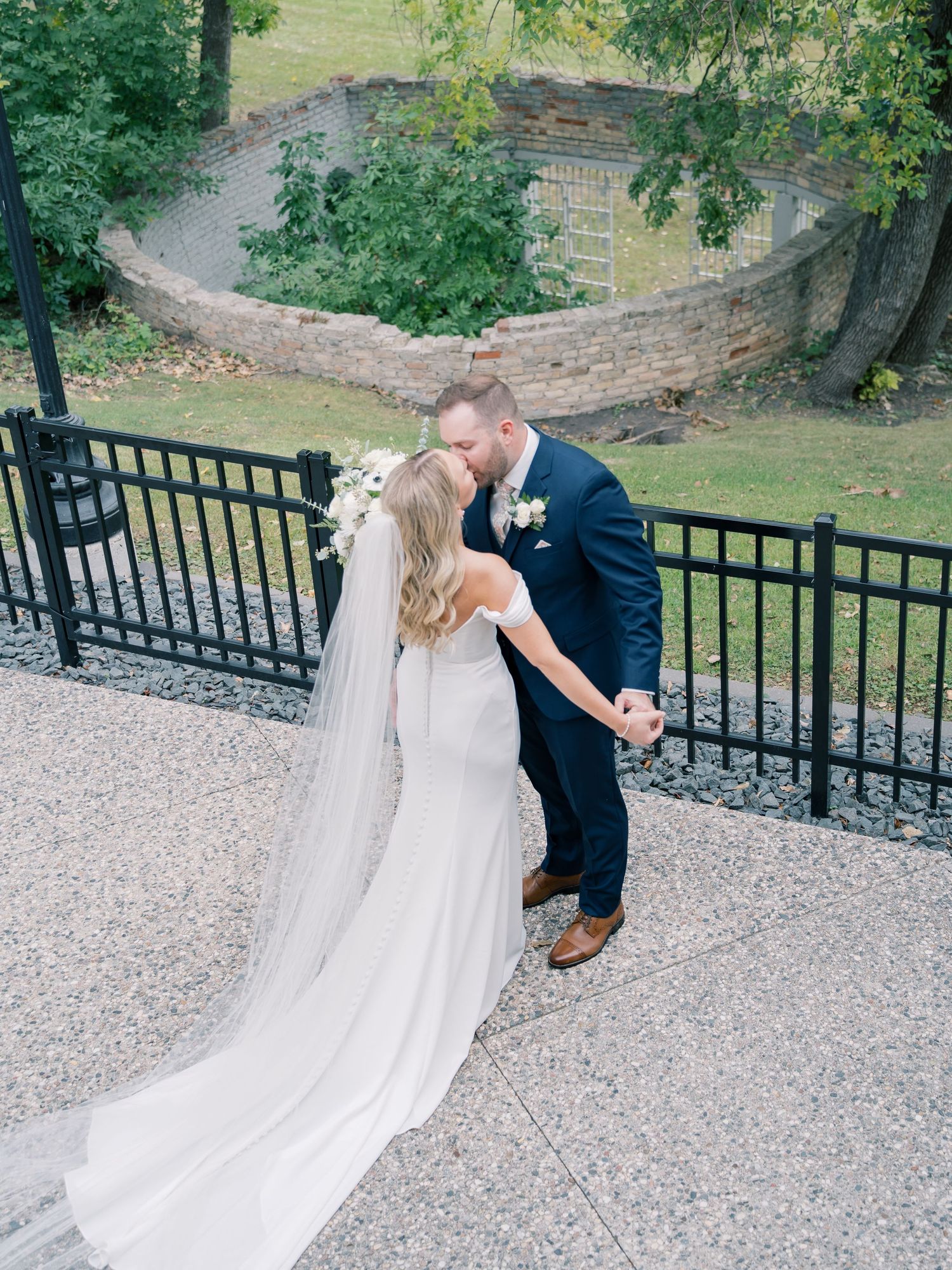 Bride and groom sharing a kiss on a paved terrace, with a black iron fence and lush greenery in the background.