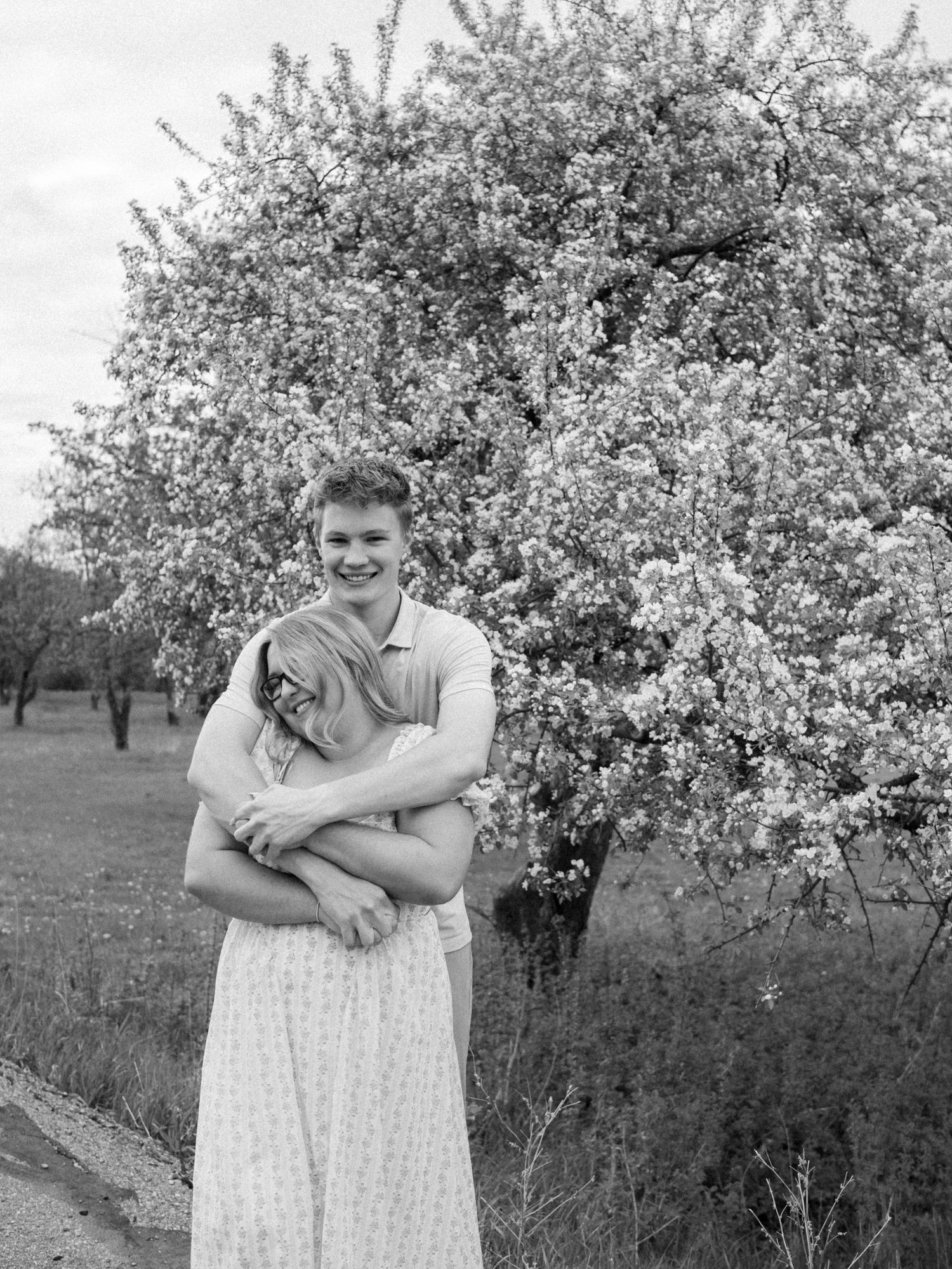 Black and white photo of a couple embracing in front of a blooming tree in a field.