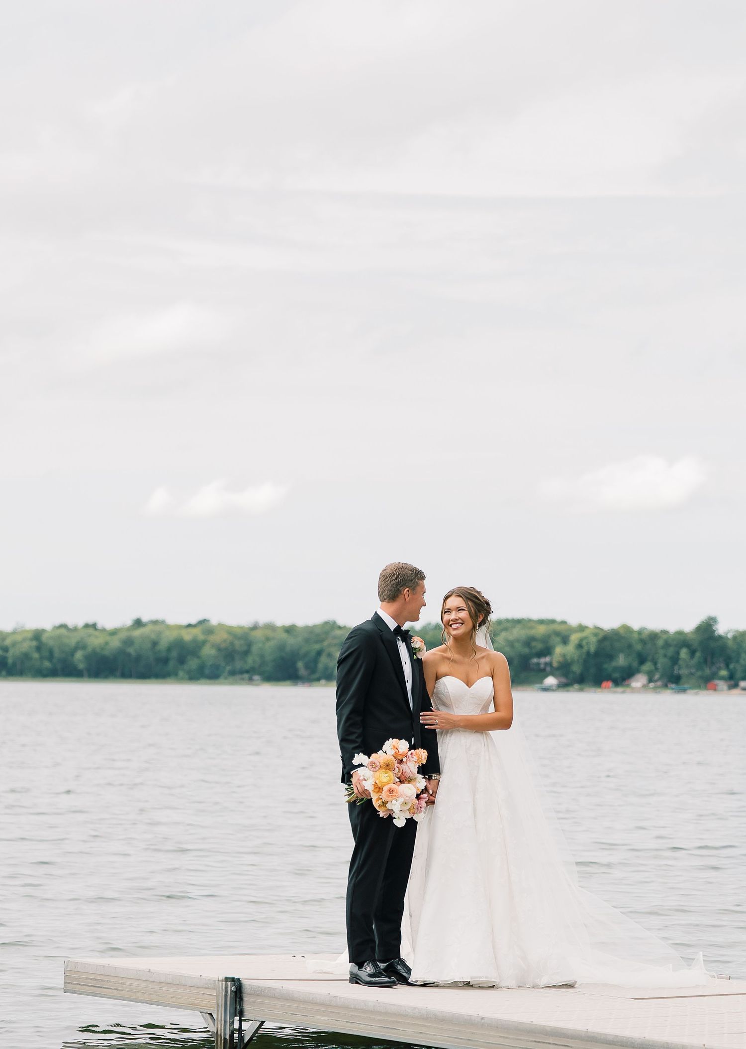 A couple in wedding attire stands on a wooden dock overlooking a serene lake surrounded by trees.