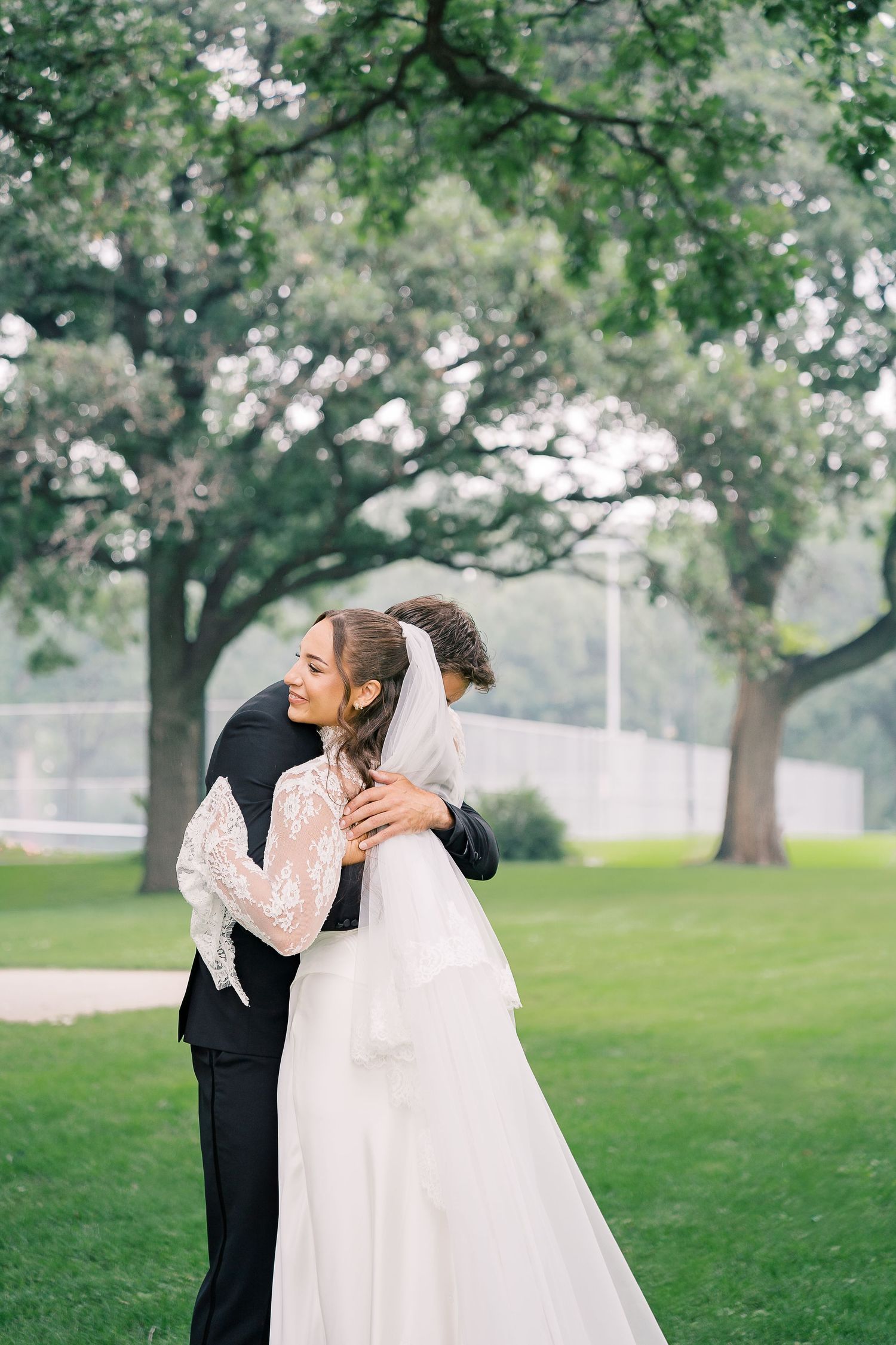 A couple embraces during their outdoor wedding ceremony under large trees in a grassy park setting.