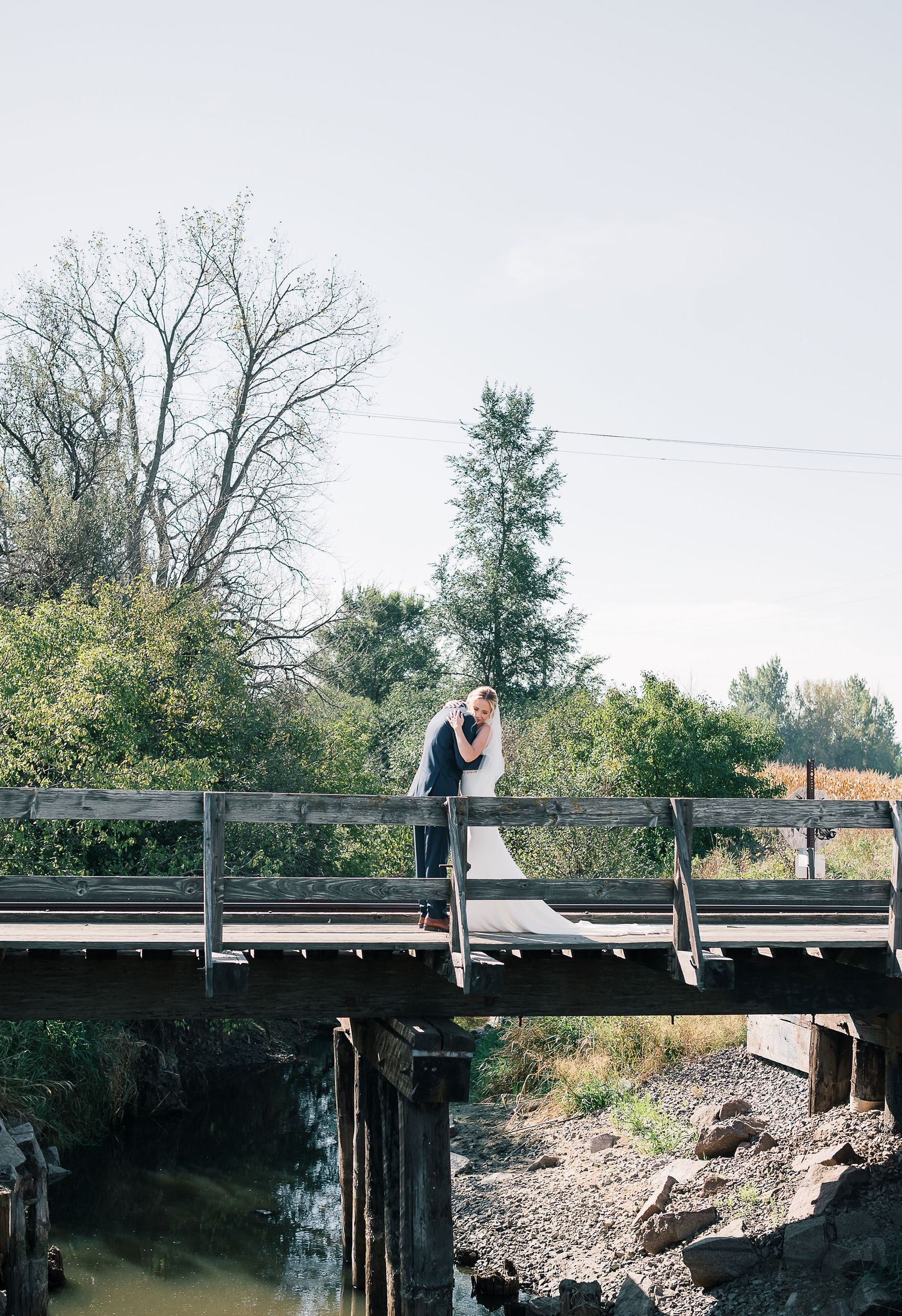 A couple stands on a rustic wooden bridge over a creek, surrounded by autumn trees in a romantic outdoor setting.