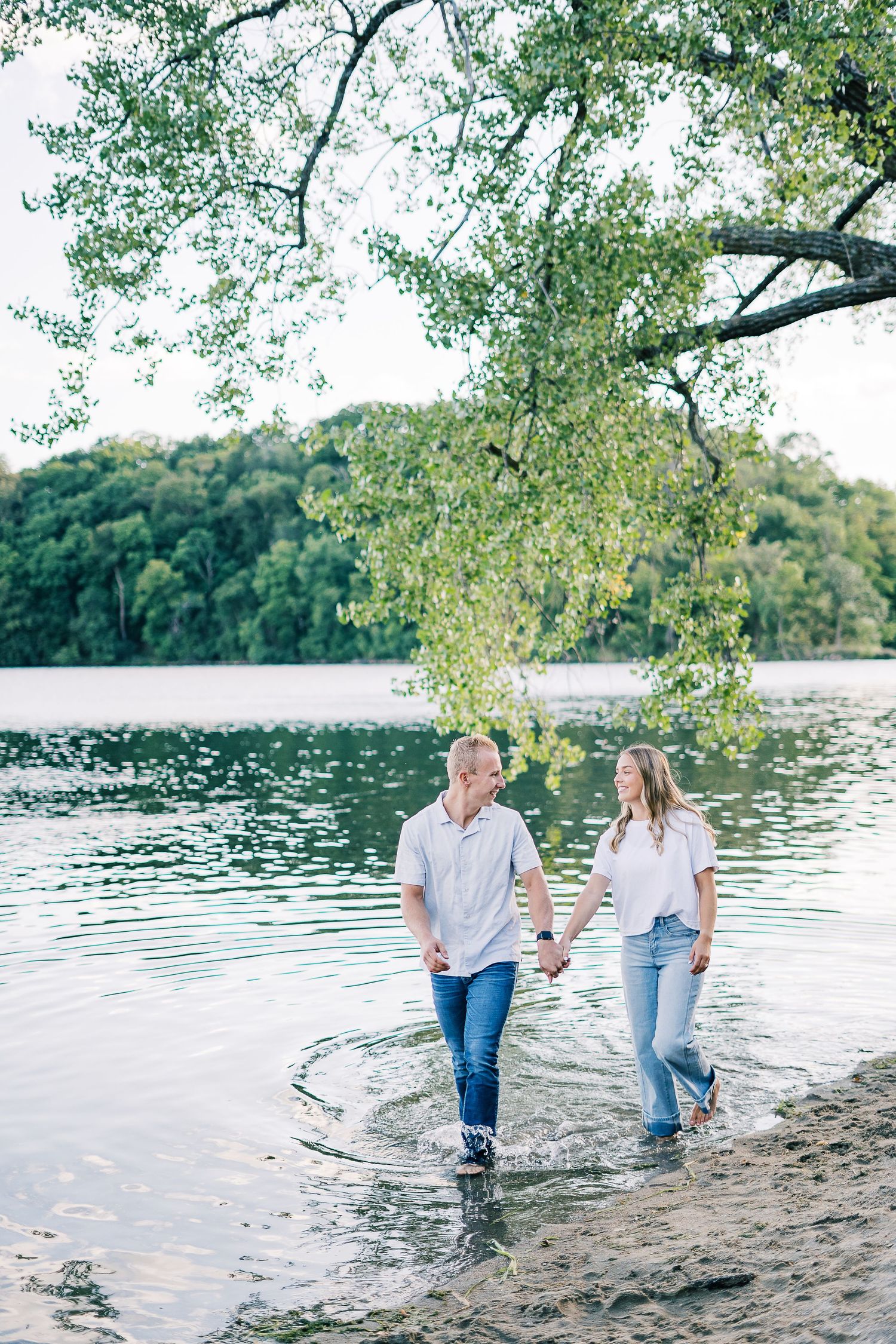 A couple walks hand in hand along a serene lakeshore beneath overhanging trees on a peaceful autumn day.