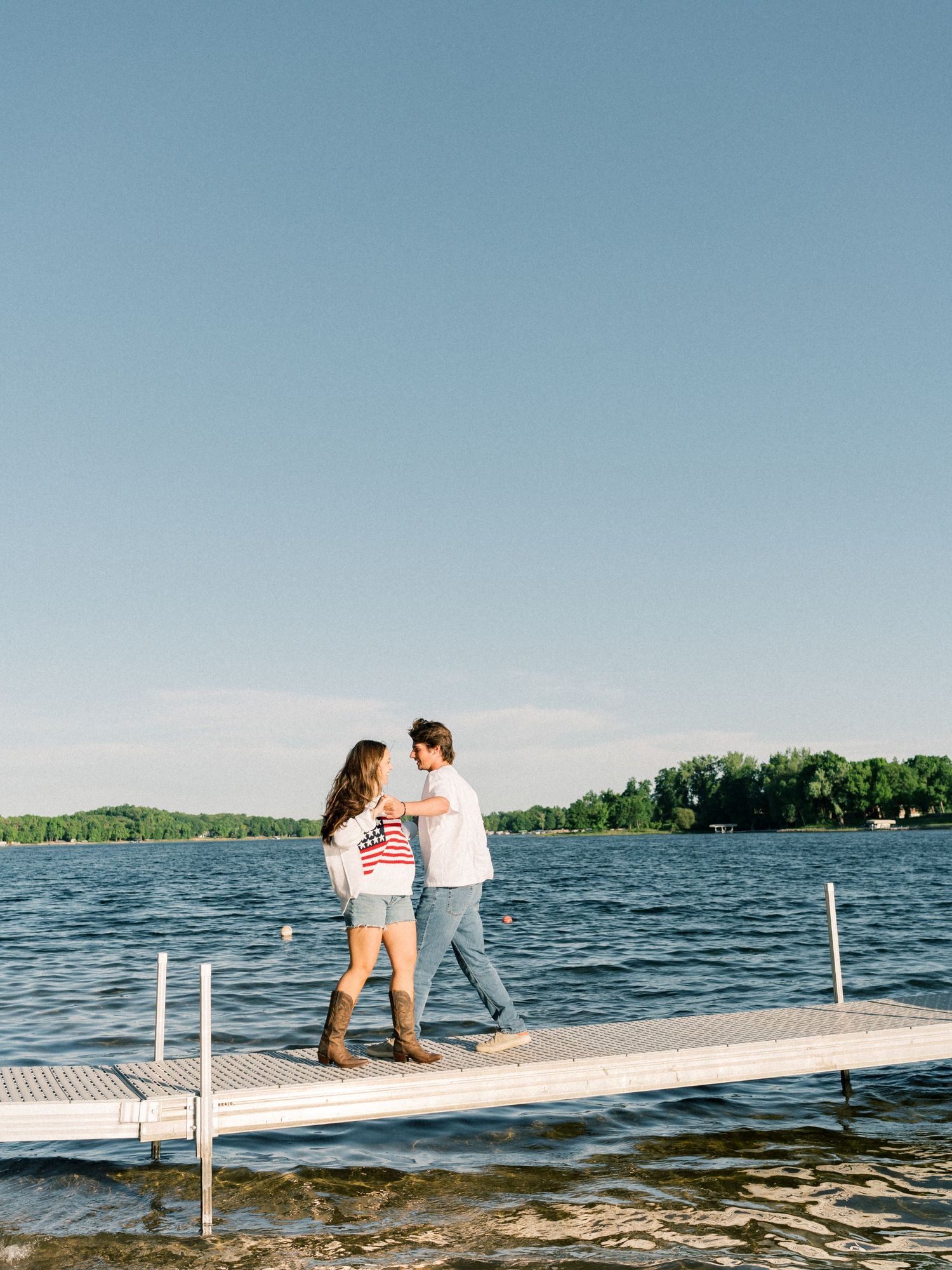 Couple walking hand in hand on a lake dock, wearing casual summer outfits, with calm blue water and green trees behind.