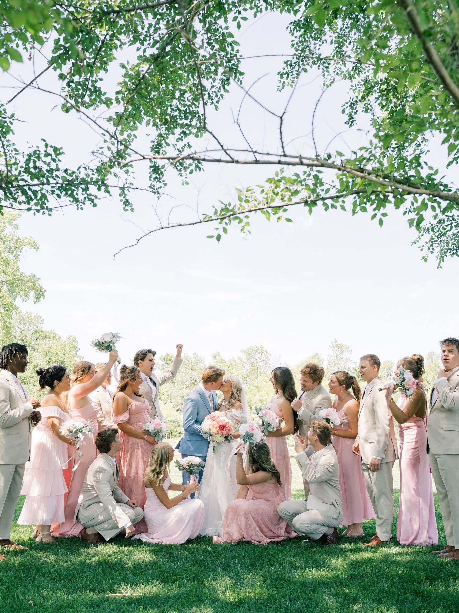 Wedding party in pink dresses and tan suits celebrates outdoors under green trees on a sunny day.