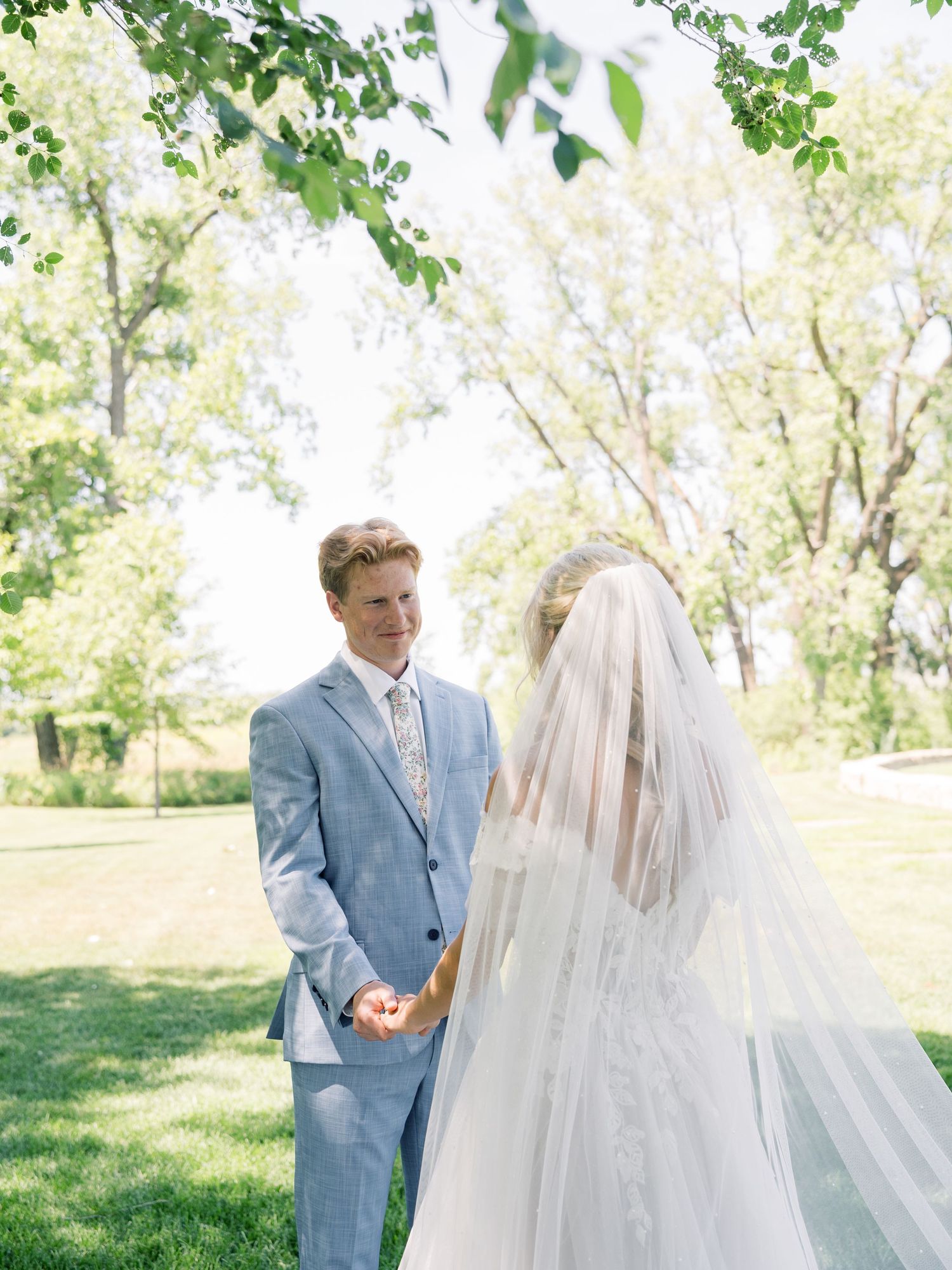 Groom in light blue suit holds bride's hand outdoors during first look, bride wearing white gown and veil.