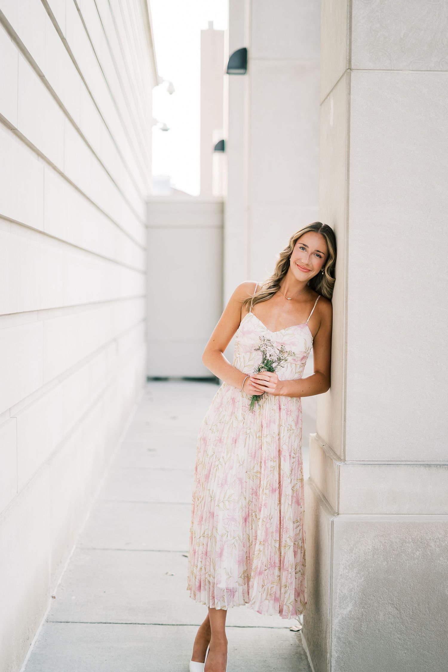 Woman in floral pleated midi dress holding small bouquet poses against white architectural wall in sunlit corridor.