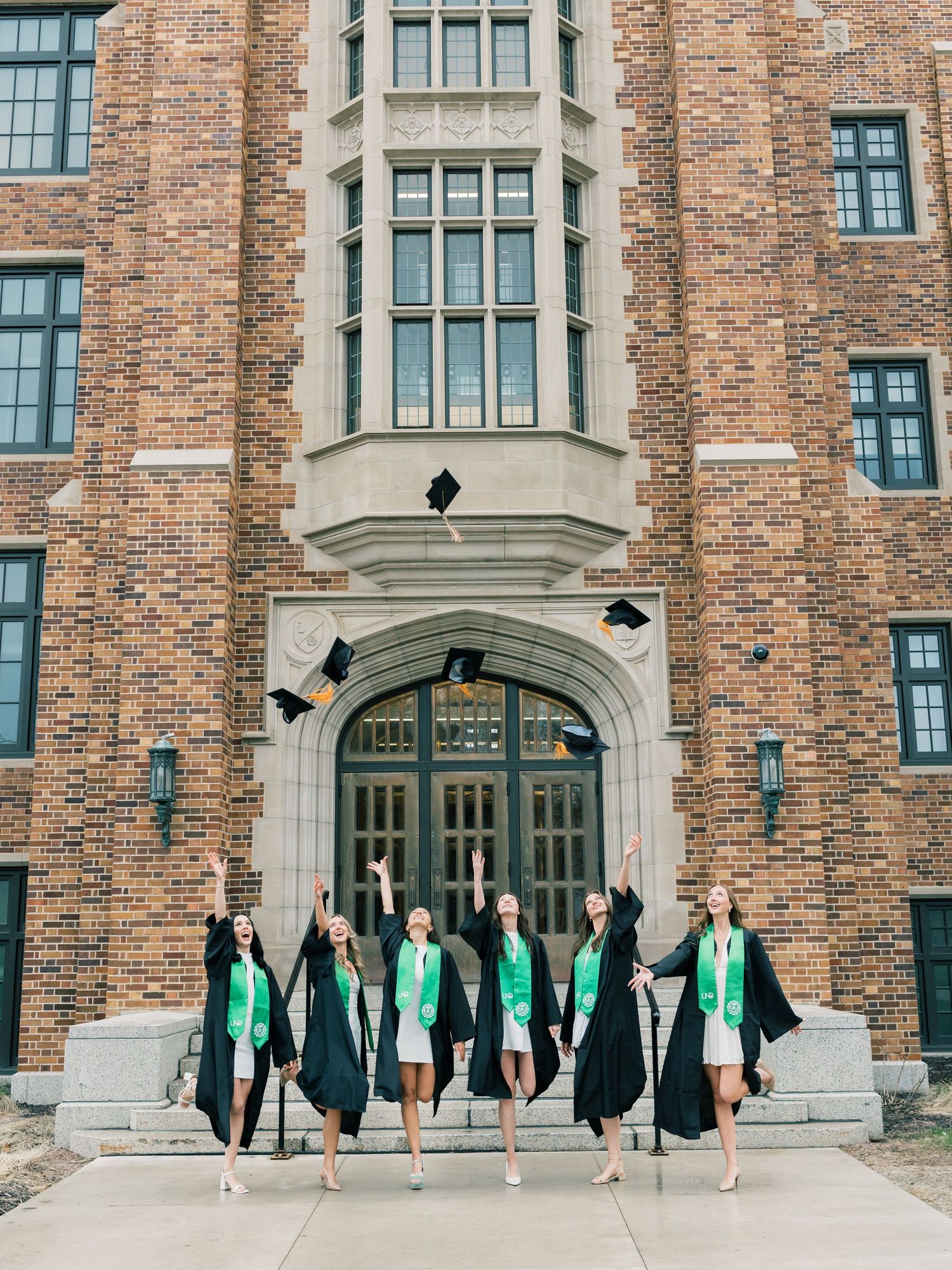 Six graduates in black robes and green stoles toss caps in front of a brick university building.