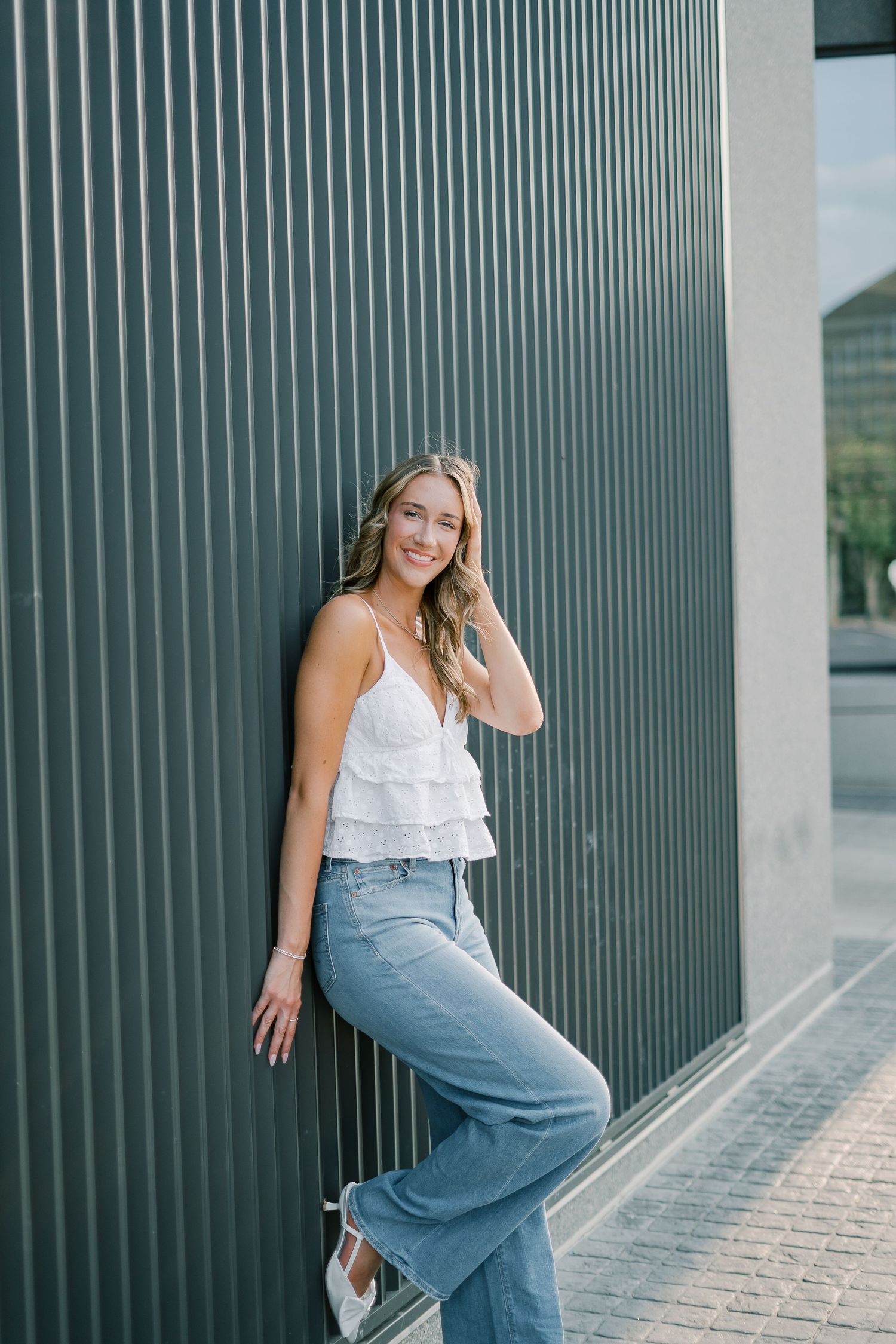 A woman in a white ruffle top and denim skirt poses against a modern green corrugated wall in an architectural setting.