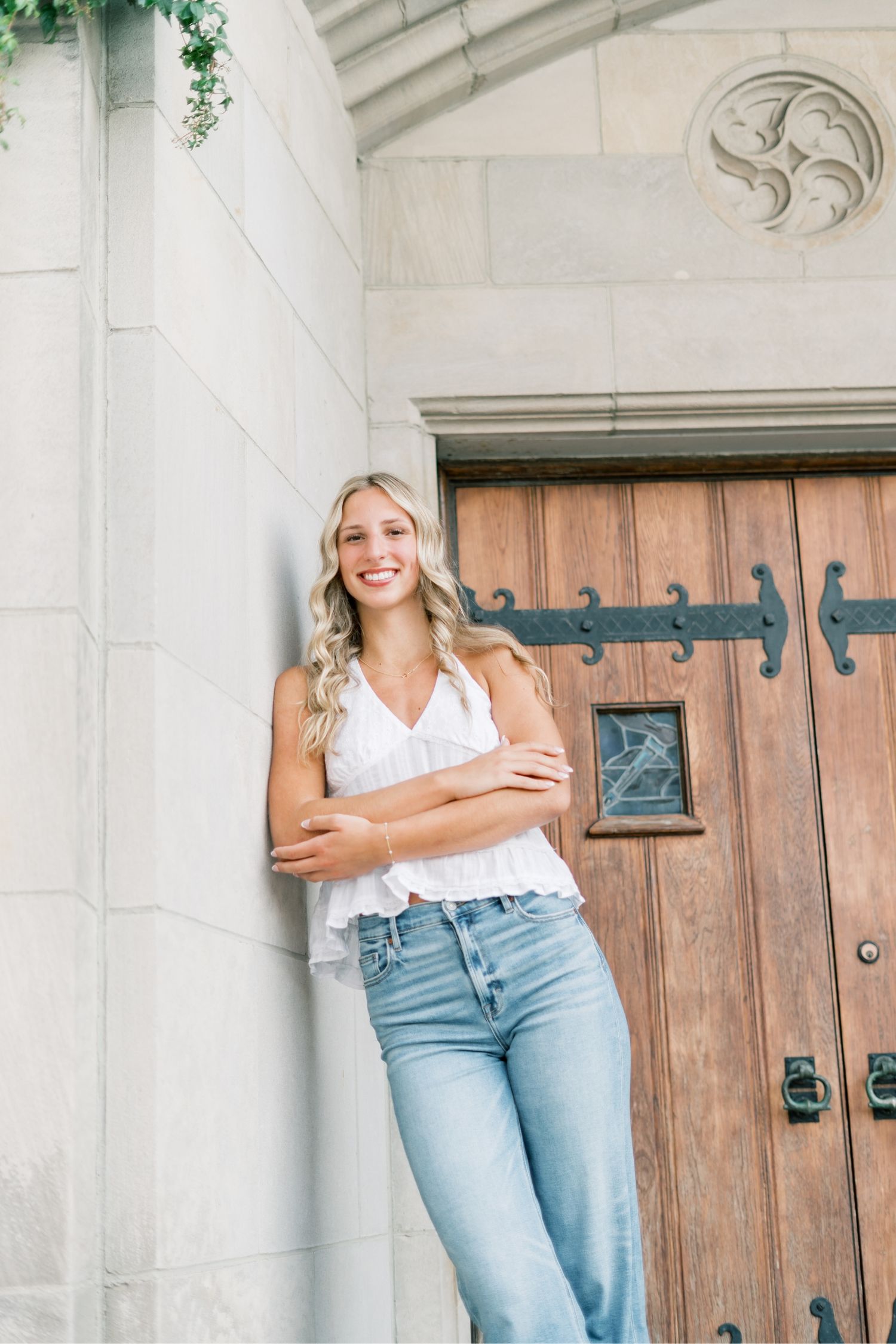 Woman in white tank top and light wash jeans poses casually against stone wall with wooden door.