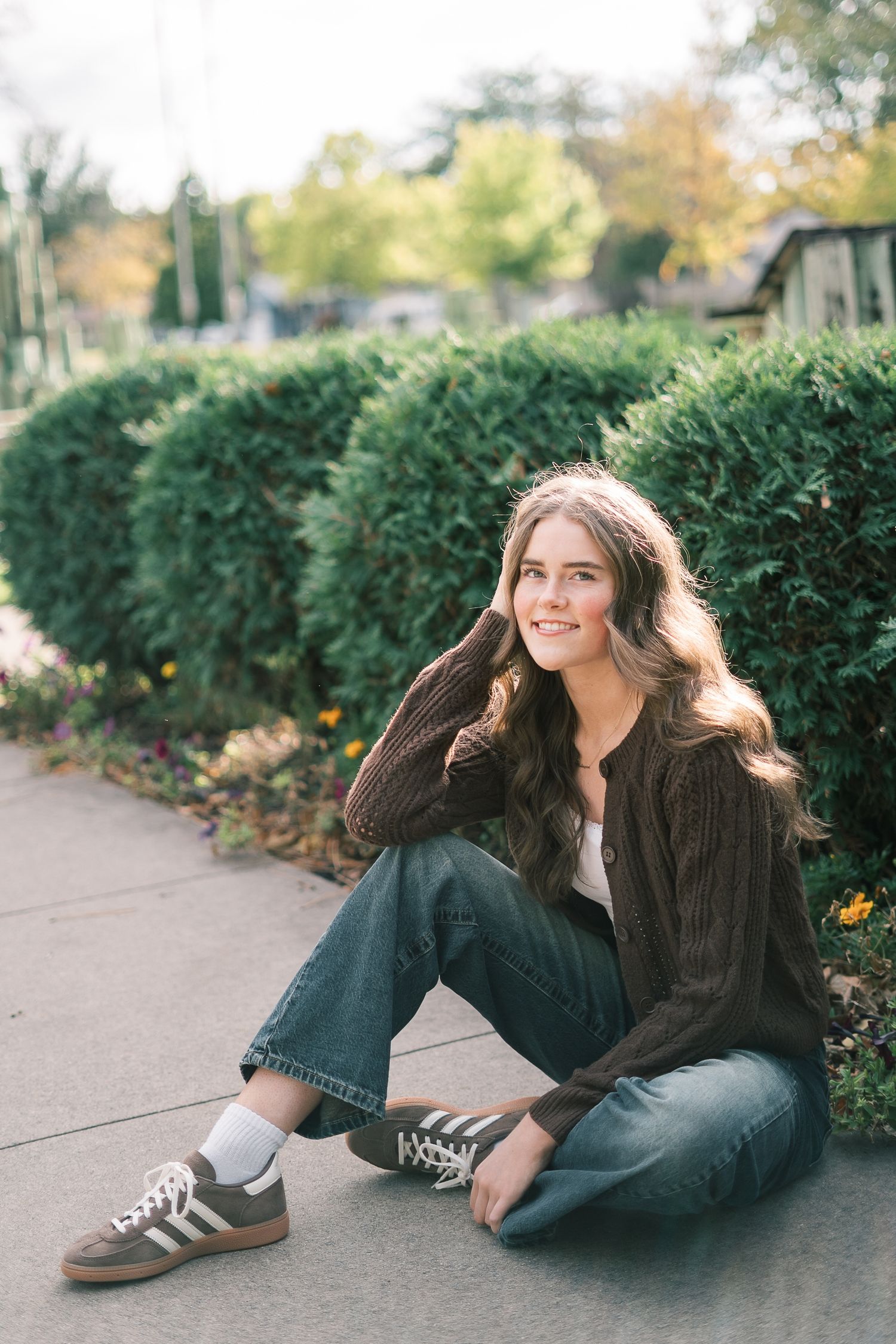 A person in a brown sweater and jeans lying casually on a sidewalk next to autumn foliage and hedges.