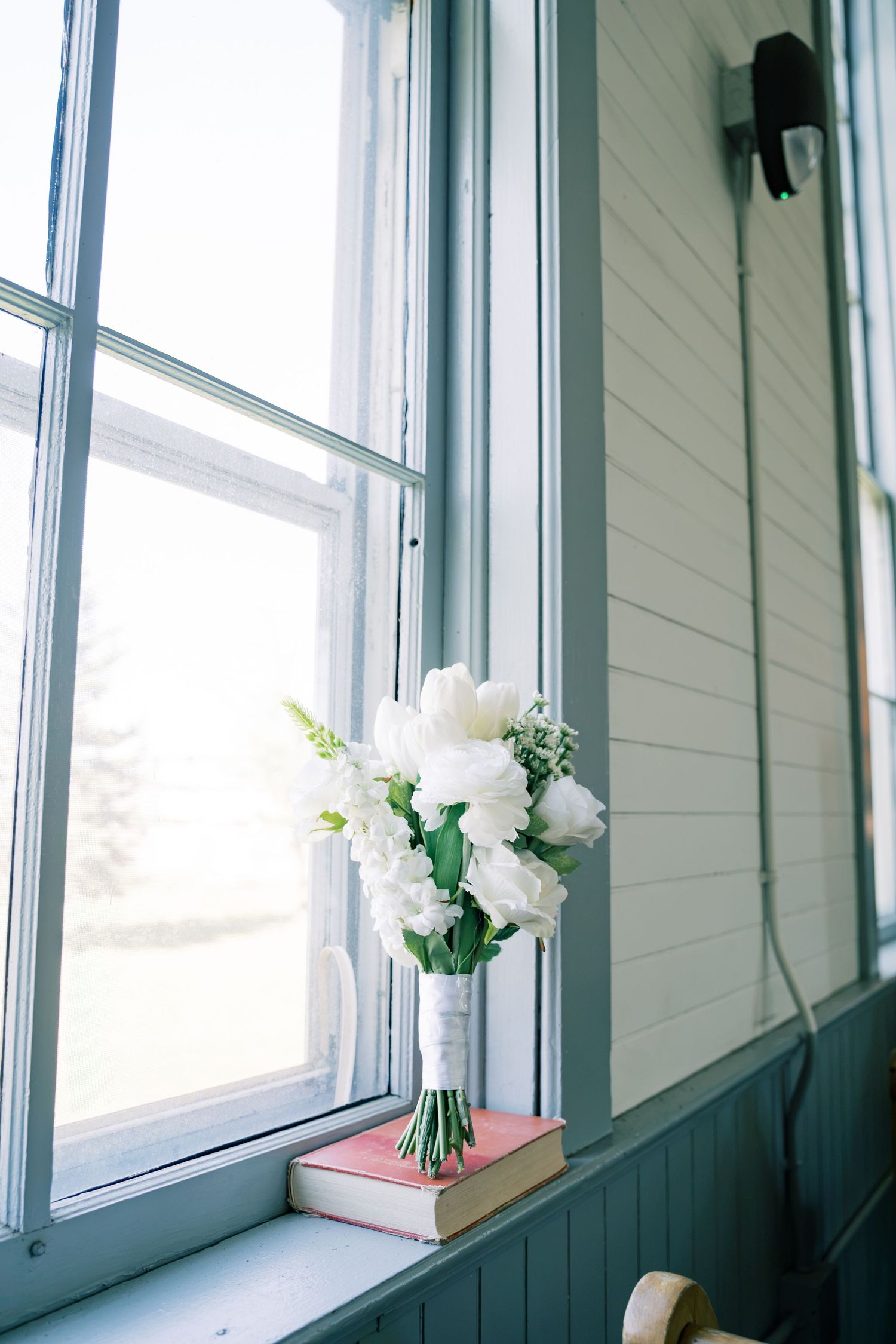 White wedding bouquet of roses and greenery resting on a pink book by a sunlit window.