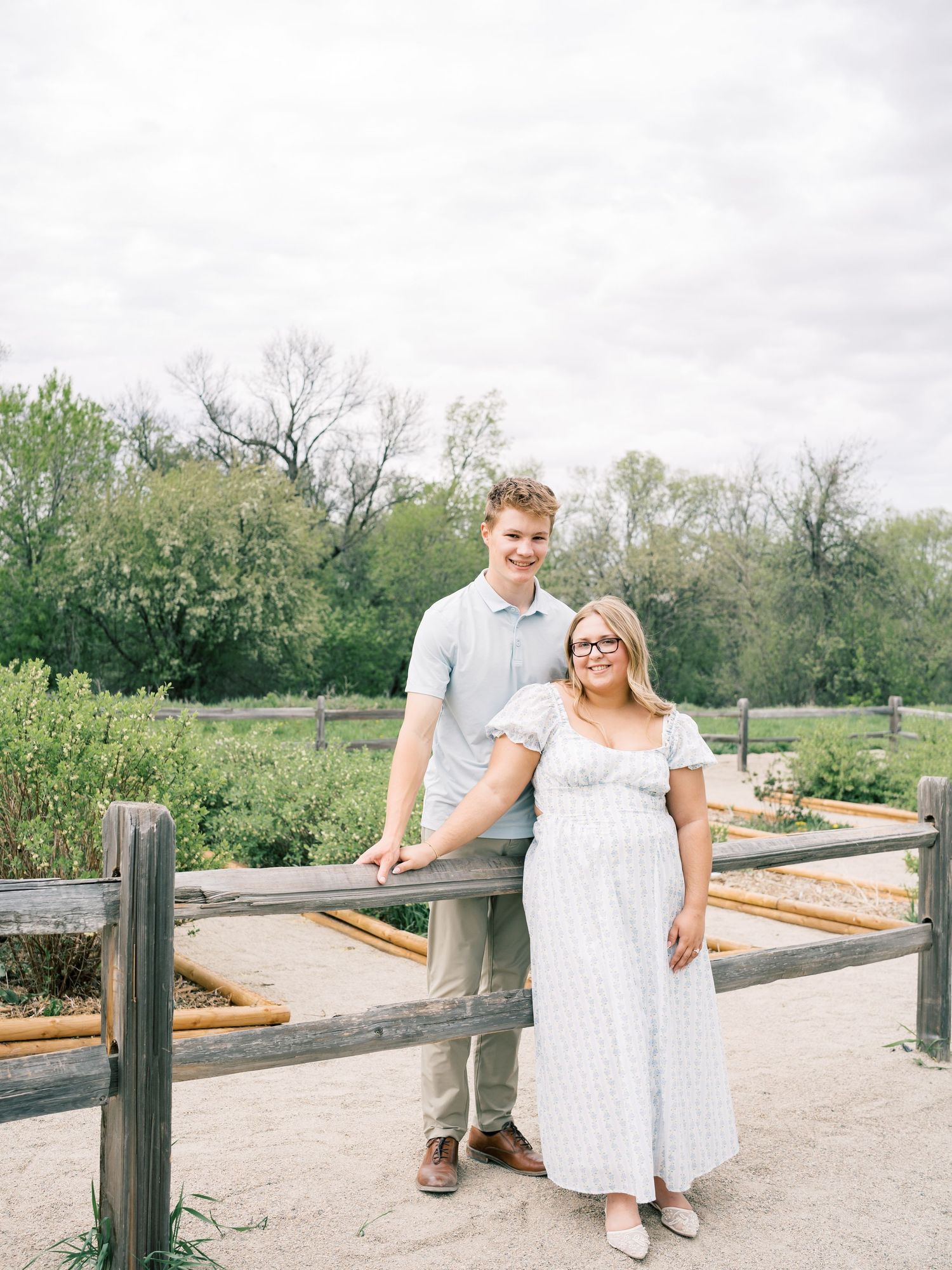A couple poses by a wooden fence in a lush garden, woman in white dress, man in light blue shirt and khakis.