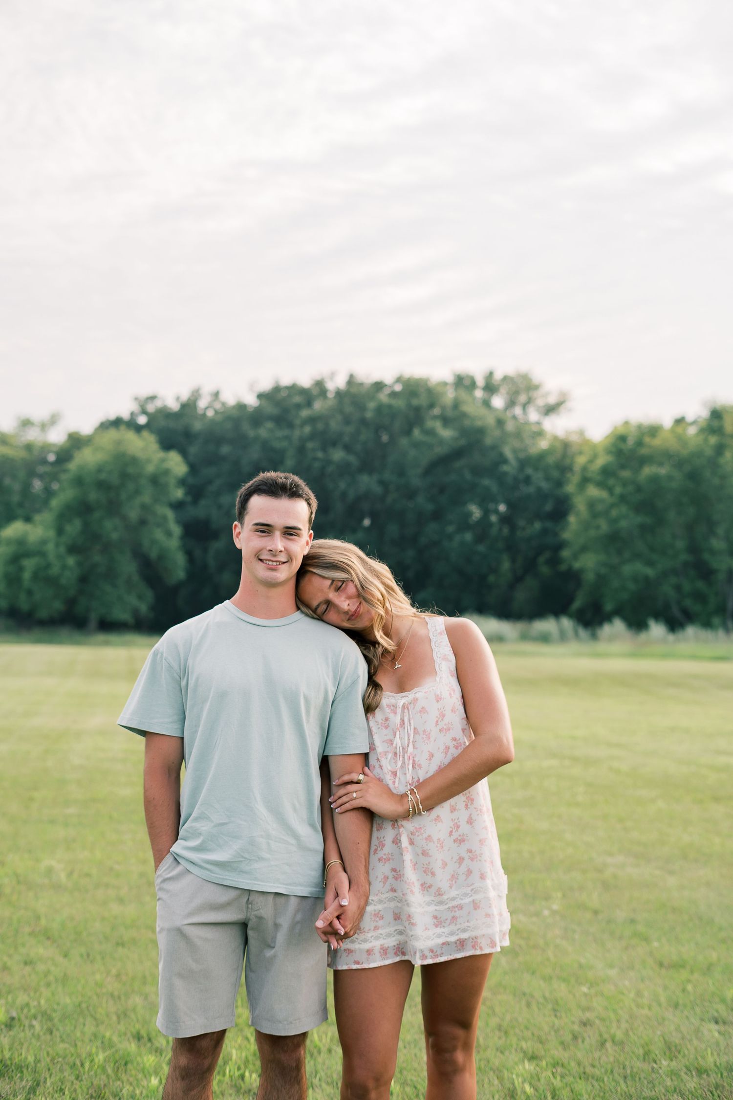 A couple stands together affectionately in a grassy field on a sunny summer day.