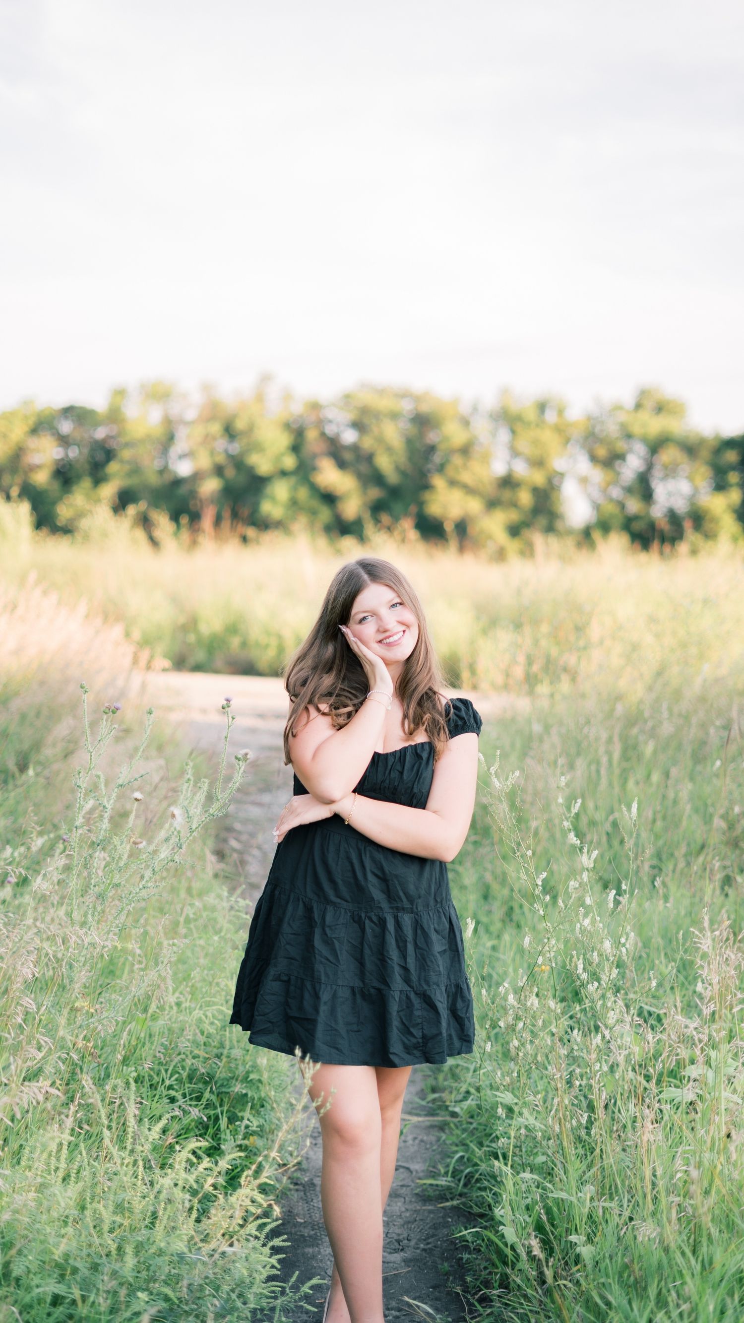 A person wearing a black dress poses playfully in a sunlit summer meadow with tall grass and wildflowers.