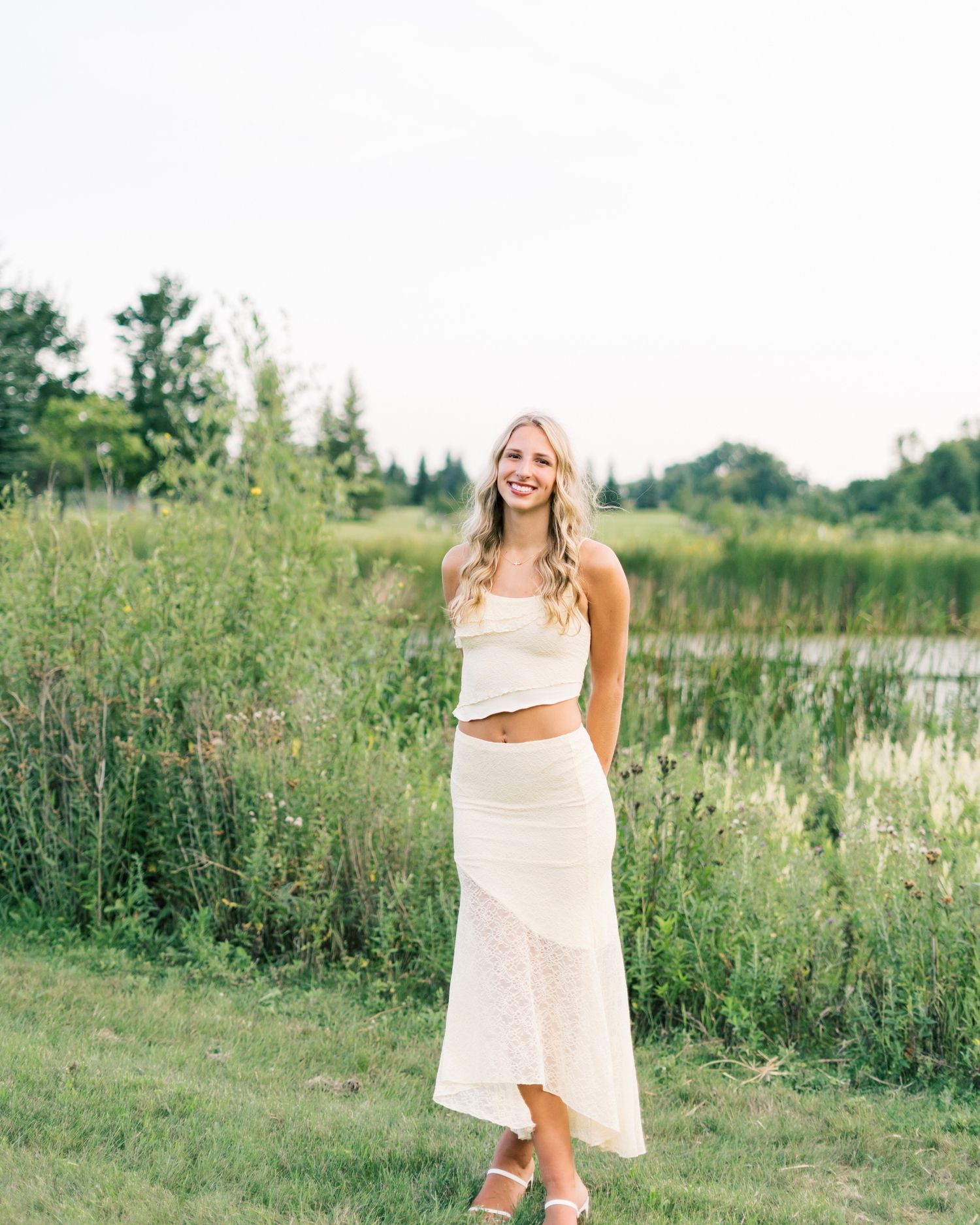Woman in a white two-piece outfit stands in a field of tall grass near a pond on a sunny summer day.