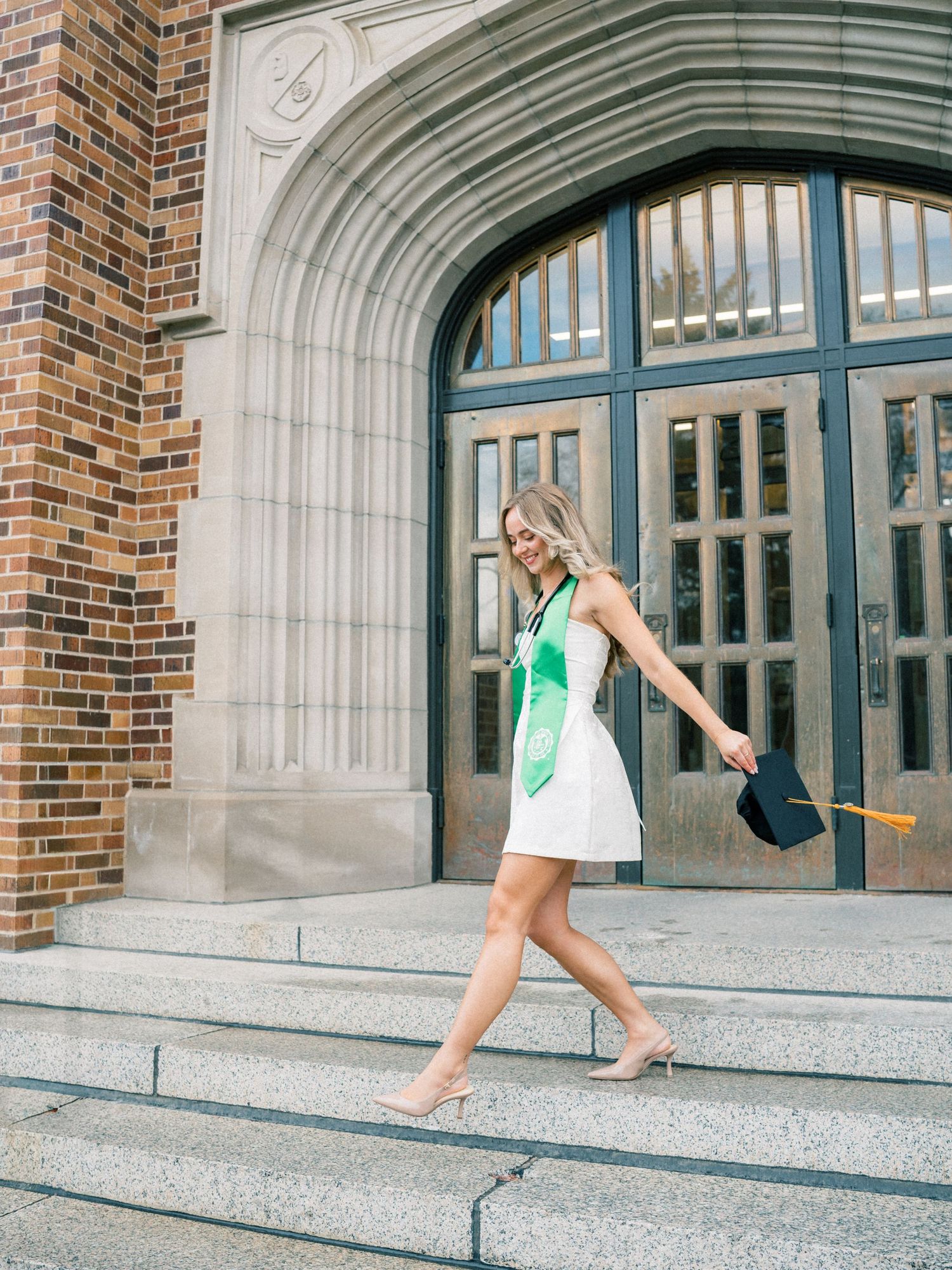 A smiling graduate in white dress with green sash walks down stone steps holding cap outside brick university building.