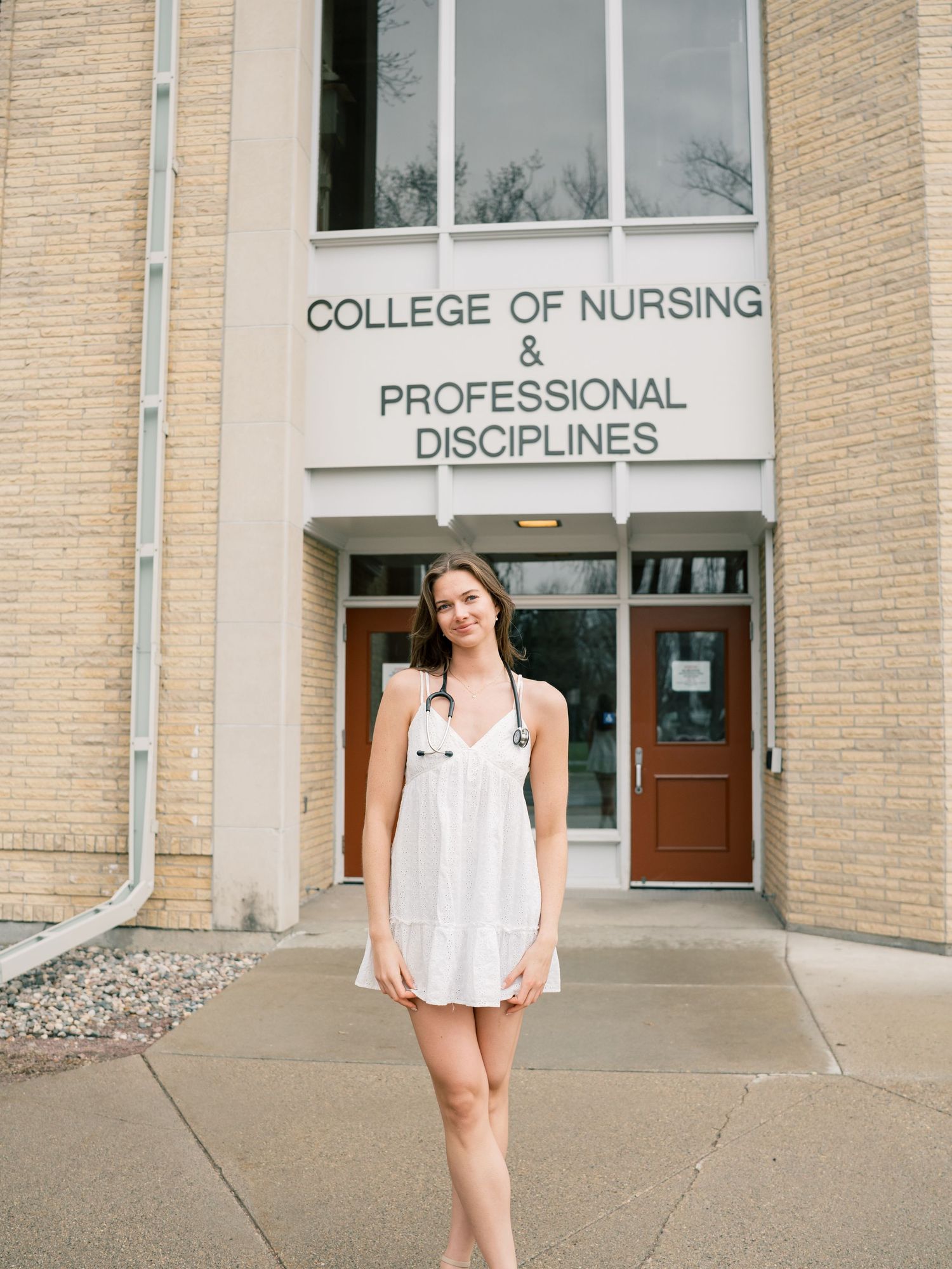 Young woman with stethoscope stands smiling in front of College of Nursing & Professional Disciplines building.