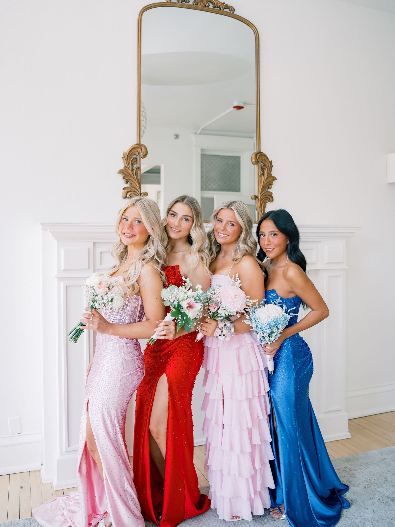 Four bridesmaids in pink, red, and blue gowns holding bouquets pose in front of a gold mirror.