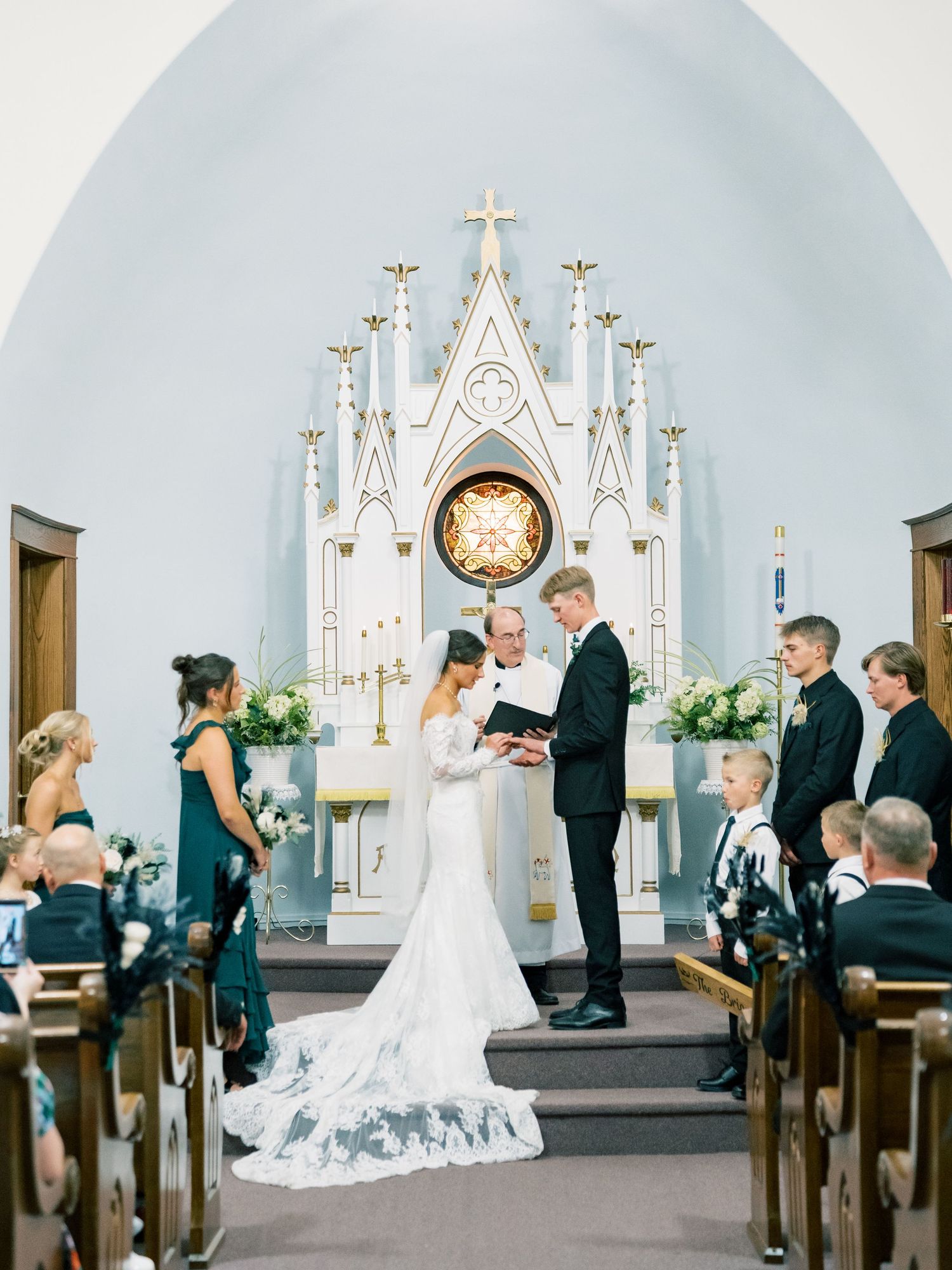 Bride and groom exchange vows at elegant white church altar with candles, surrounded by wedding party in emerald gowns.