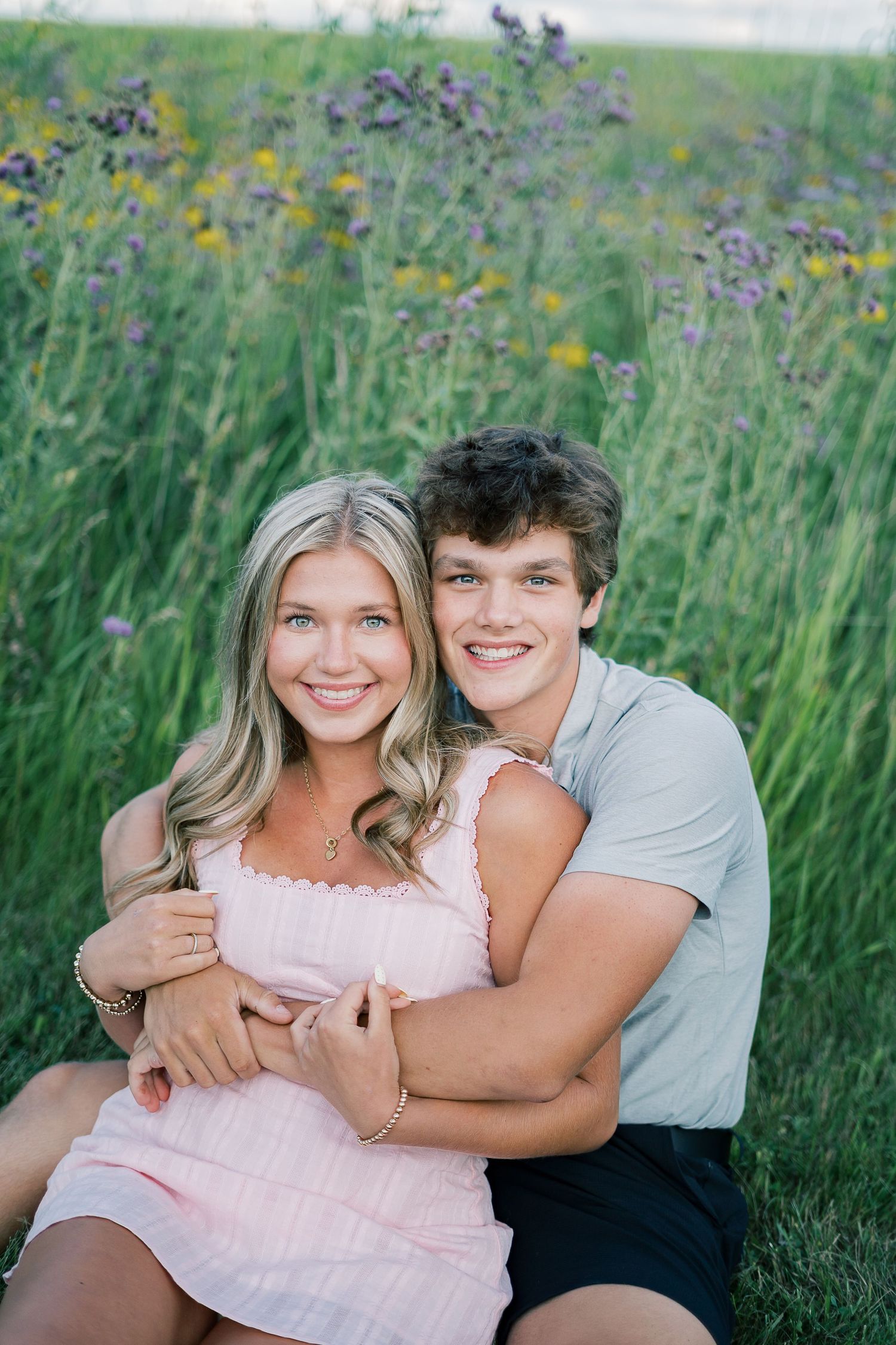 A couple embraces joyfully while sitting together in a field of tall wildflowers and grass on a sunny day.