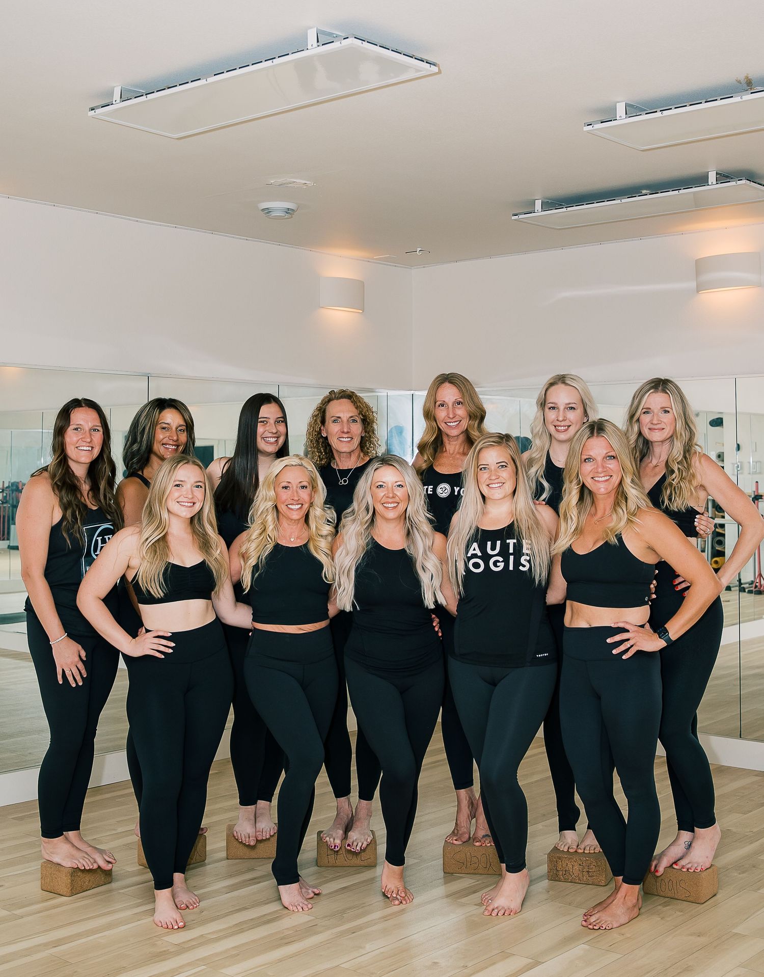 A group of dancers in matching black outfits pose together in a mirrored dance studio.