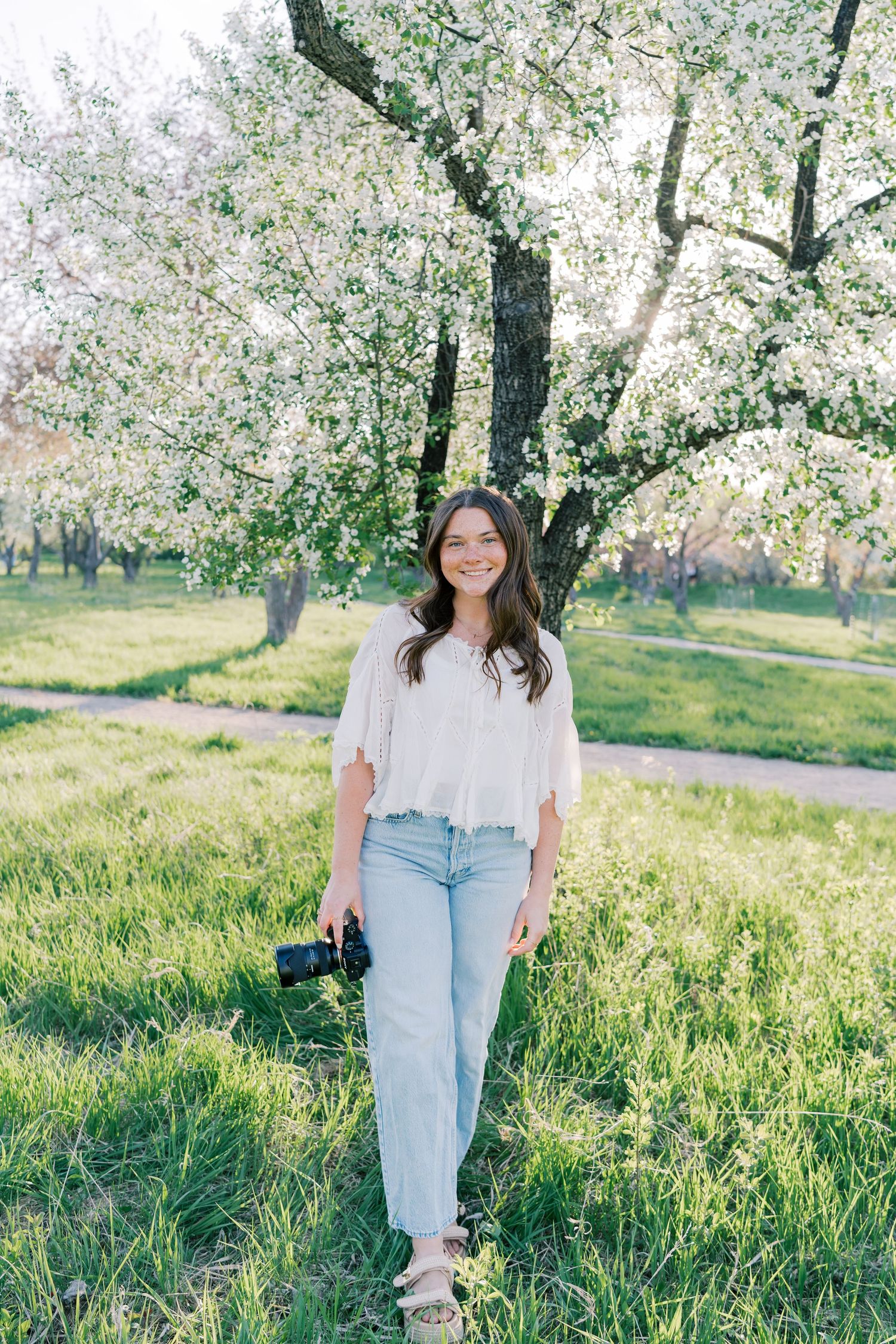 Young woman smiling in a blooming orchard, holding a camera, wearing jeans and white top on green grass.