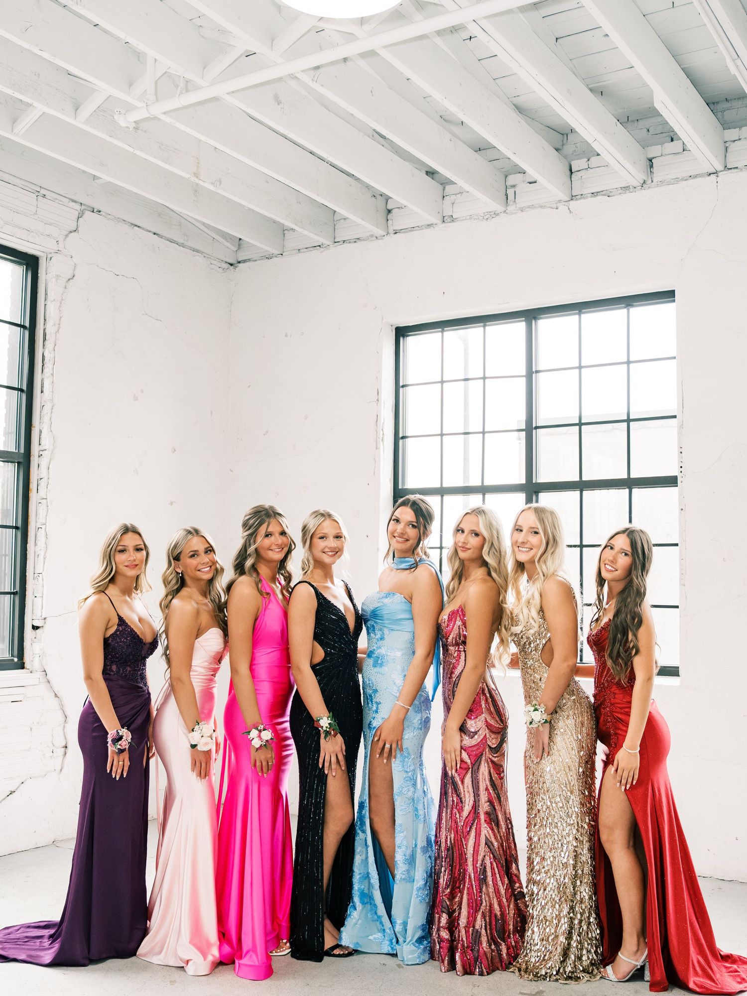 Eight young women in colorful formal prom gowns pose together in a bright white industrial-style venue.