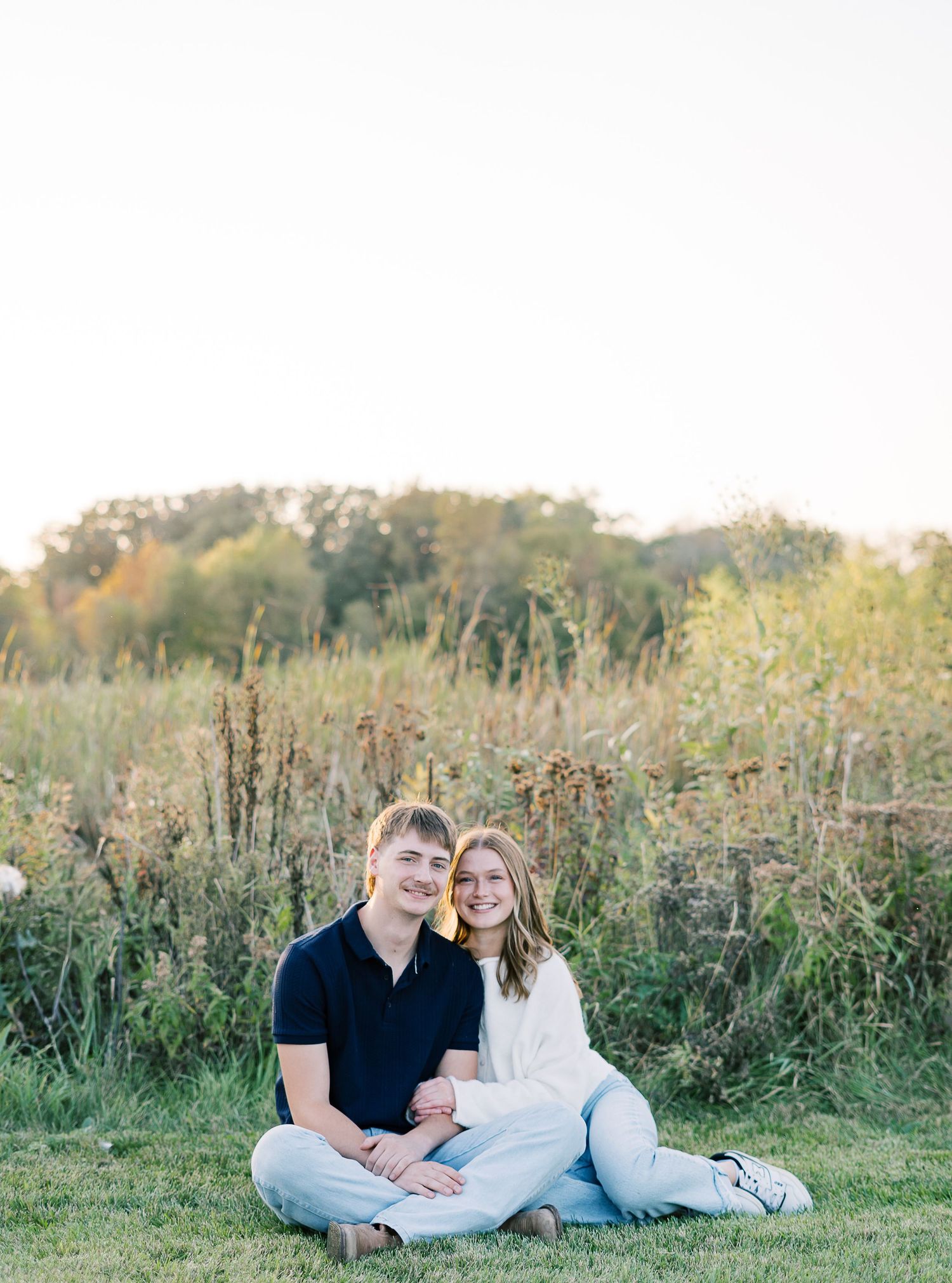 A couple sits together in a grassy meadow during golden hour with wildflowers and natural foliage surrounding them.