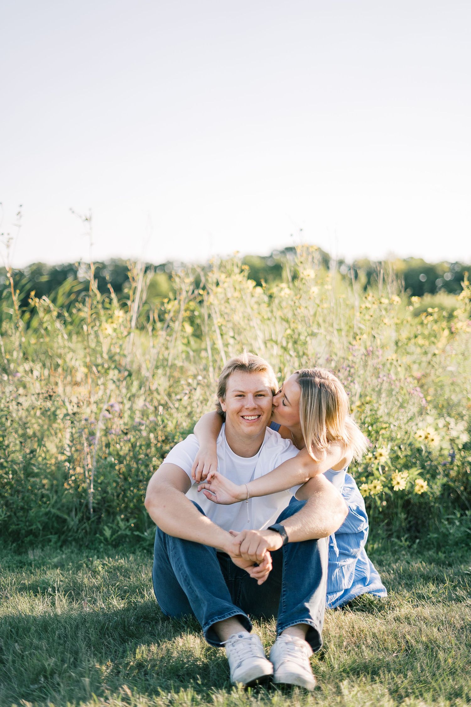 A couple shares playful moments together while sitting in a sunlit meadow with wildflowers.