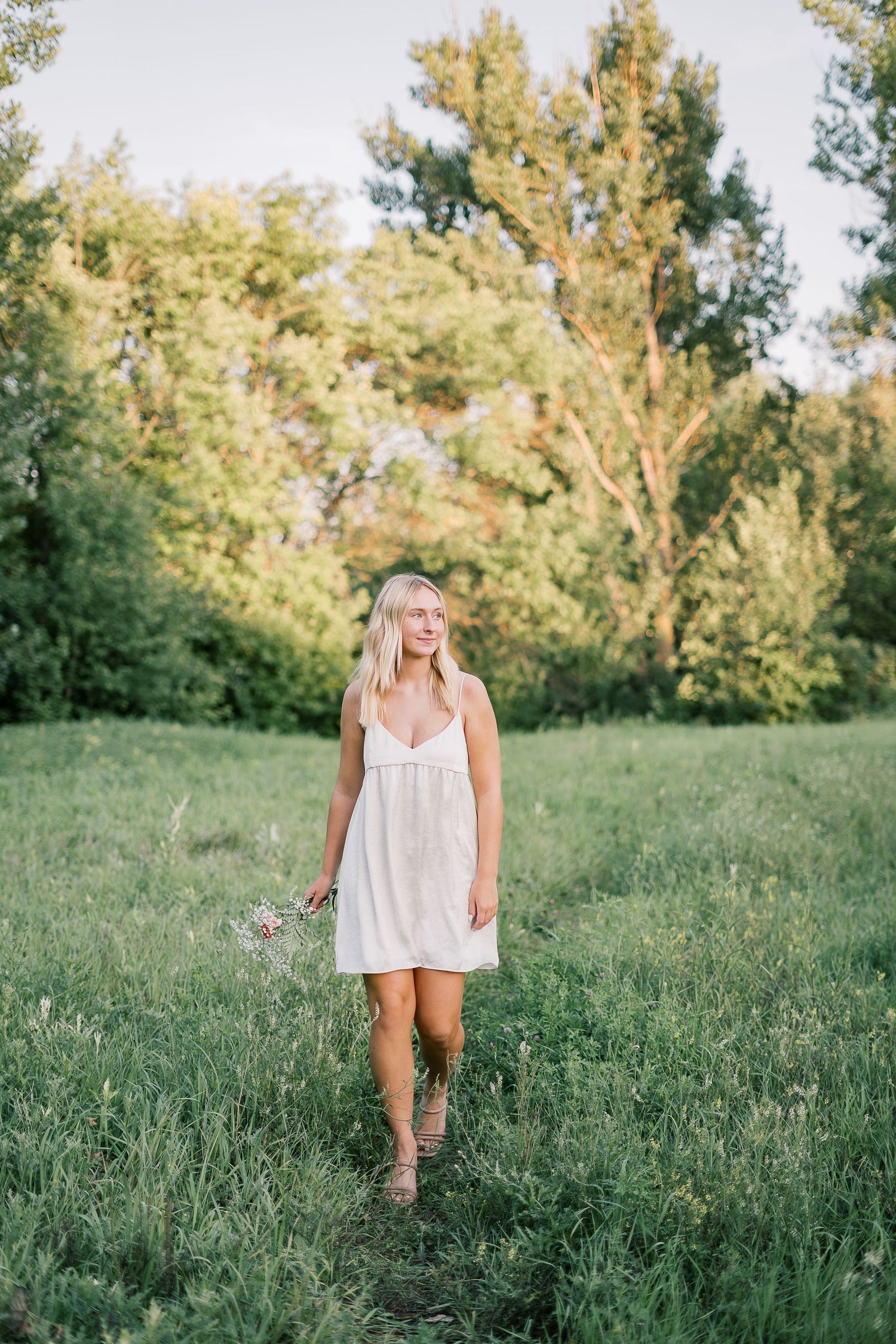 A person in a white dress walks through a sunlit meadow with tall grass and trees in the background.