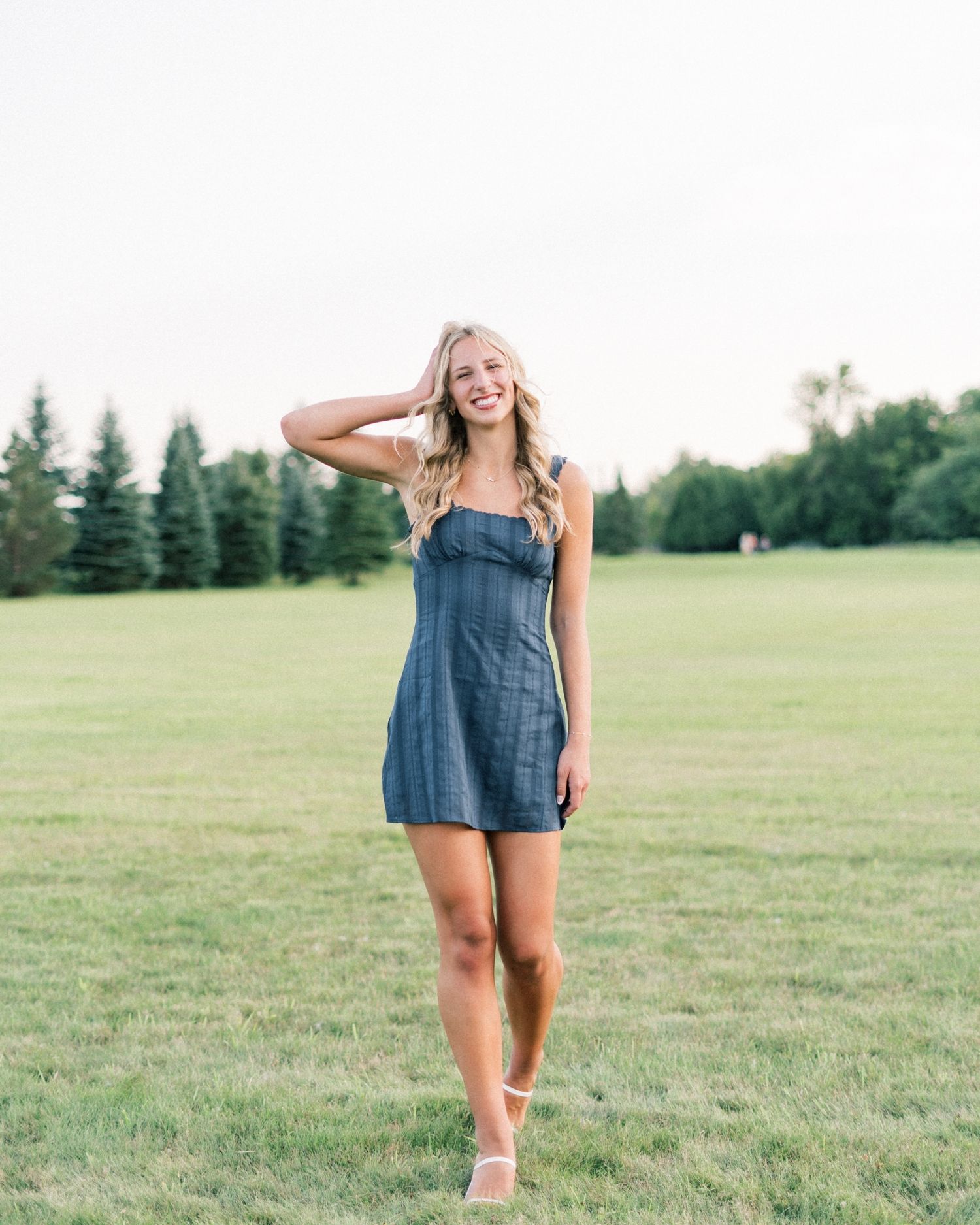 A person in a denim dress walks and poses playfully in a sunny green field with trees in the background.