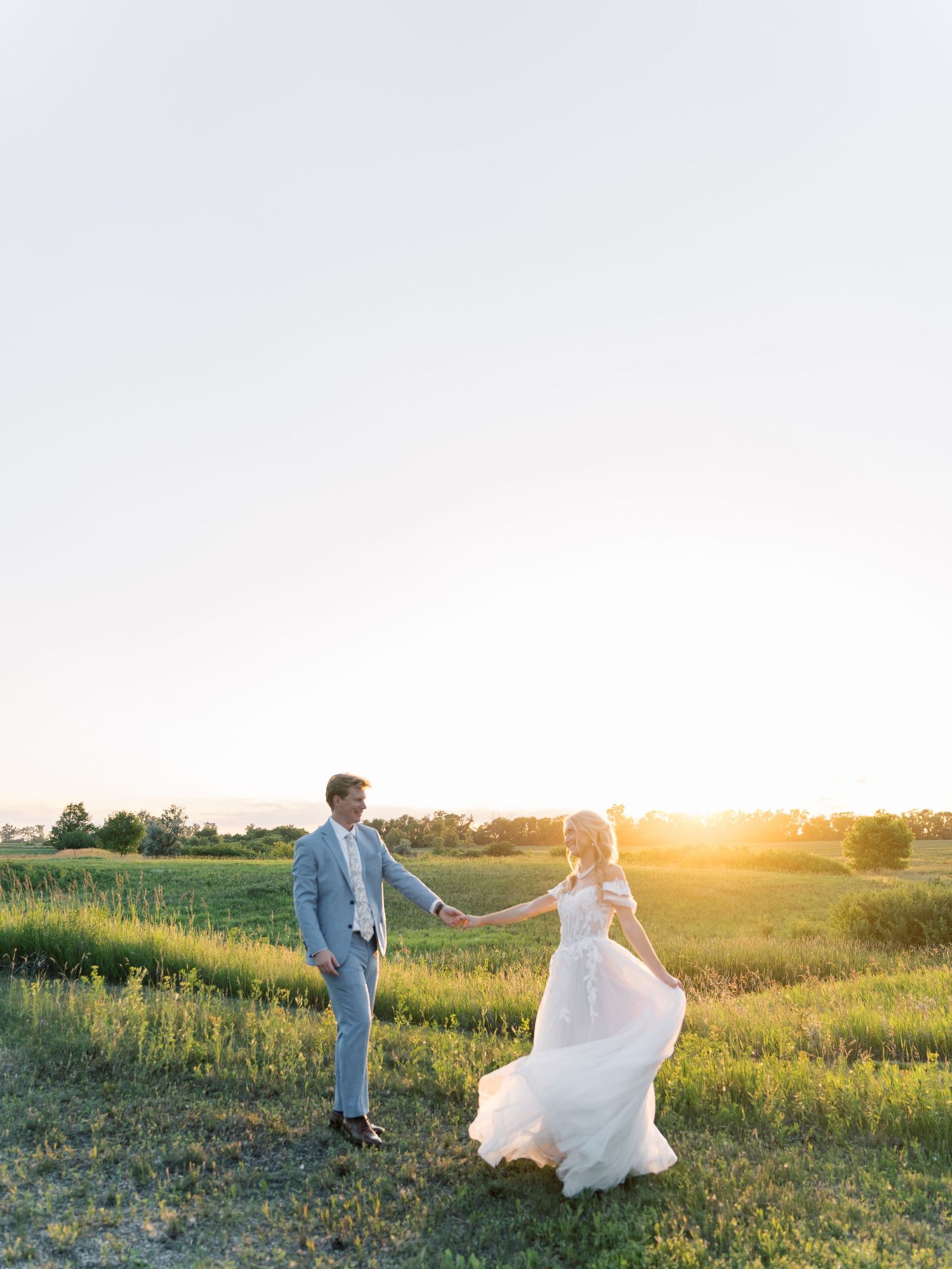 Bride and groom holding hands in a golden sunlit field at sunset, bride's white gown flowing in the breeze.