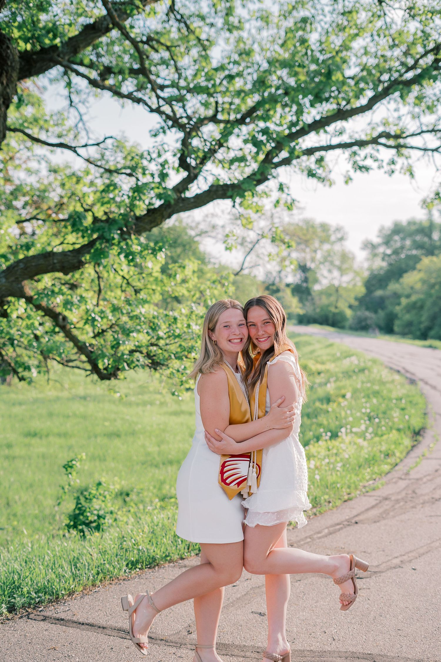 Two graduates in white dresses and gold stoles embrace while posing on a winding path.