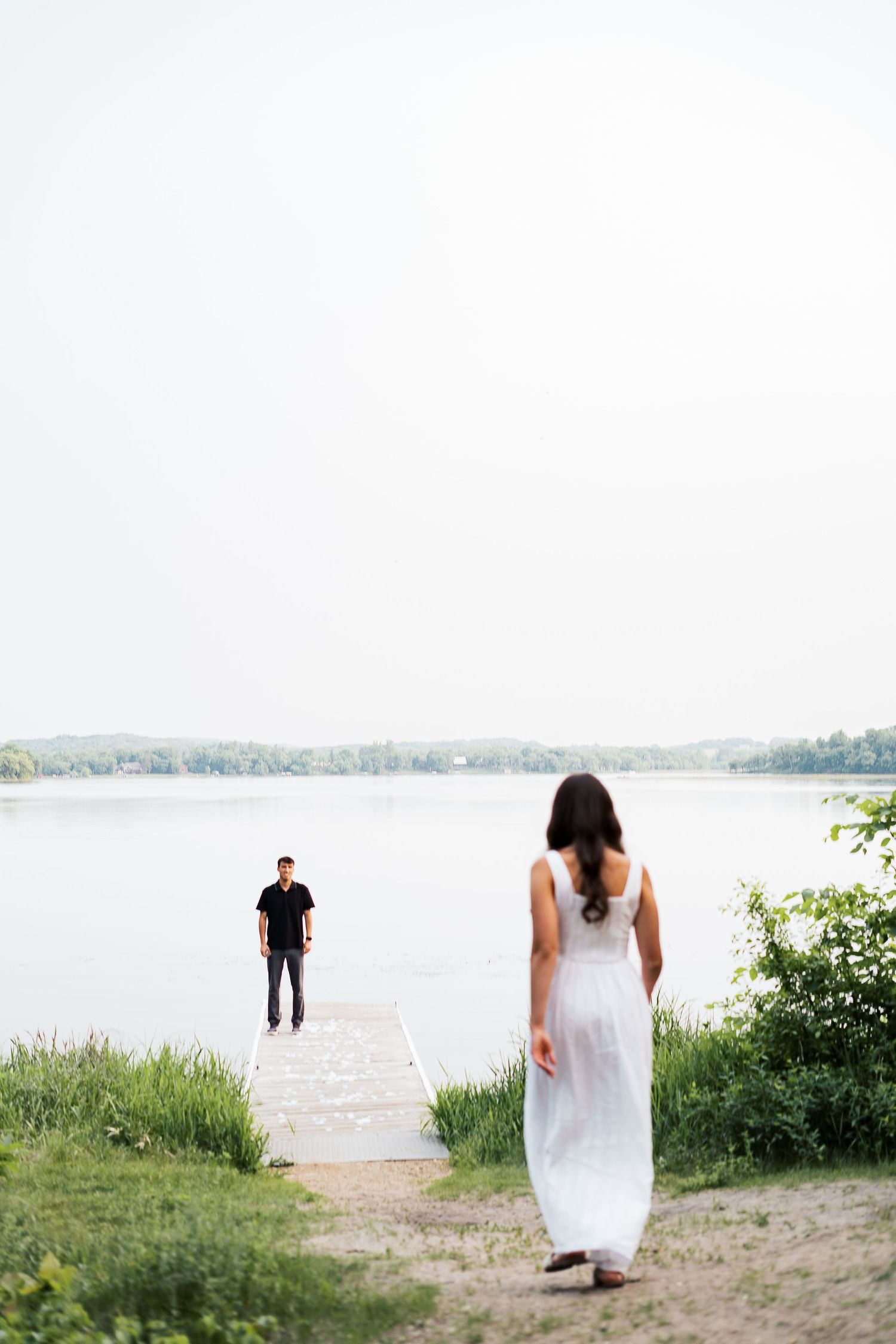 A couple stands on a lakeside path with one in a flowing white dress and another approaching on the dock.