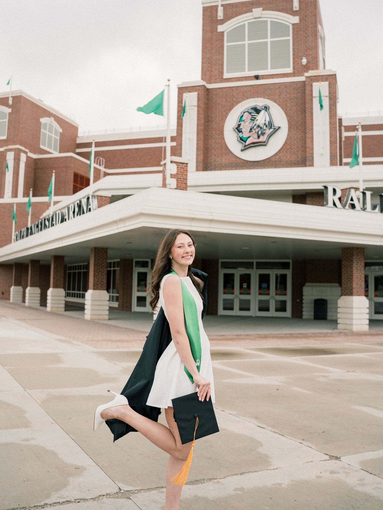 A graduate in a black cap and gown walks joyfully outside a brick university building with green flags.