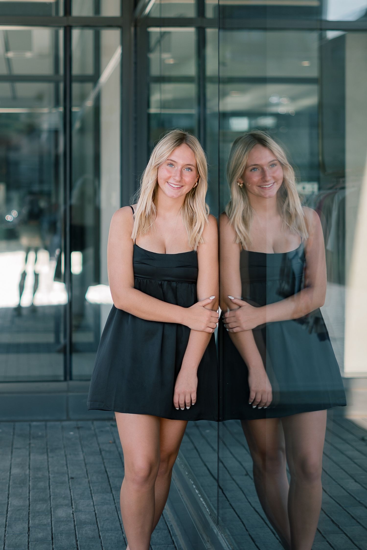 A figure in a black dress poses against a glass wall with their reflection creating a mirrored effect.