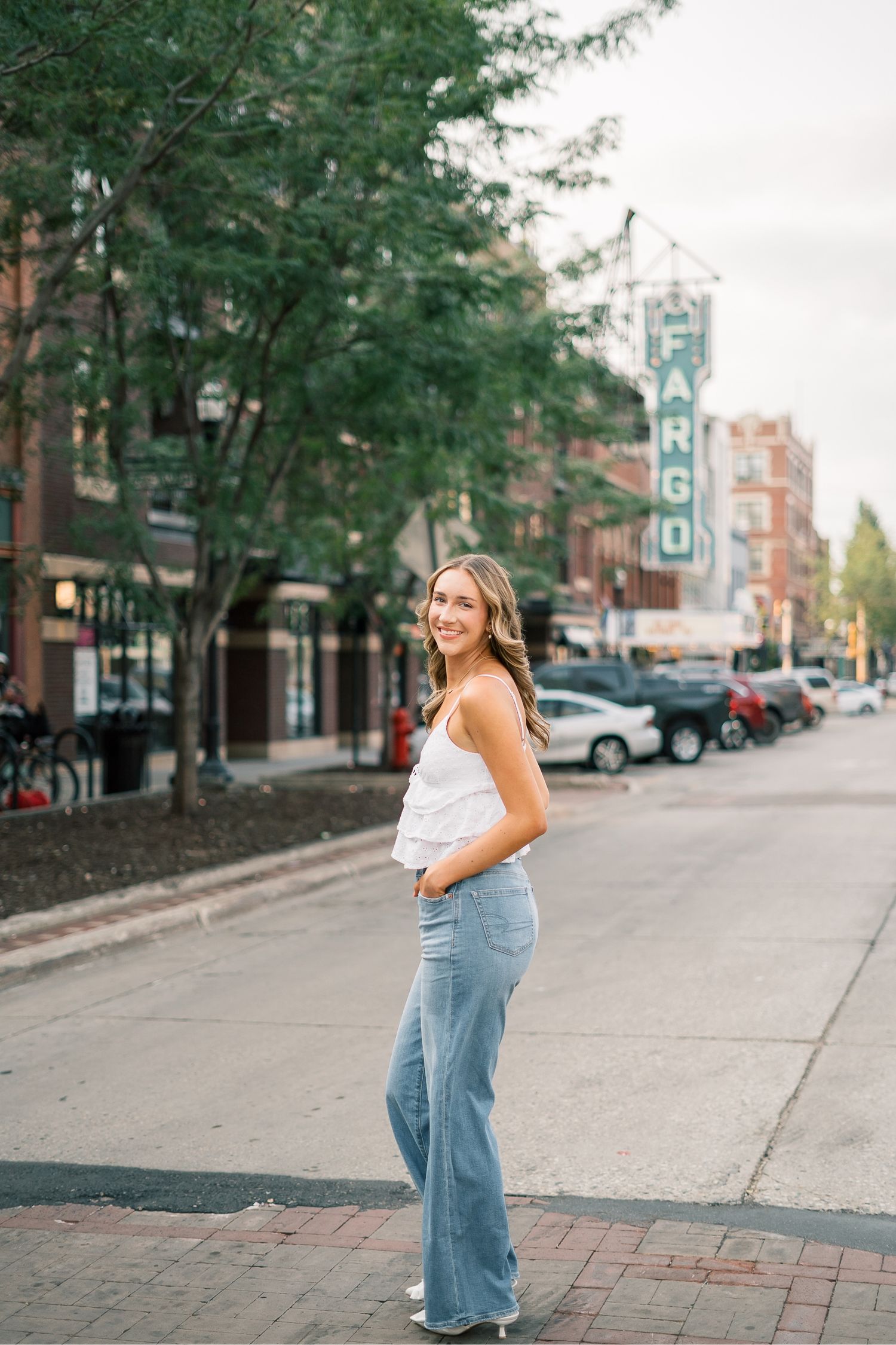 A person in a white top and flared jeans stands on a city street with brick buildings and trees in the background.