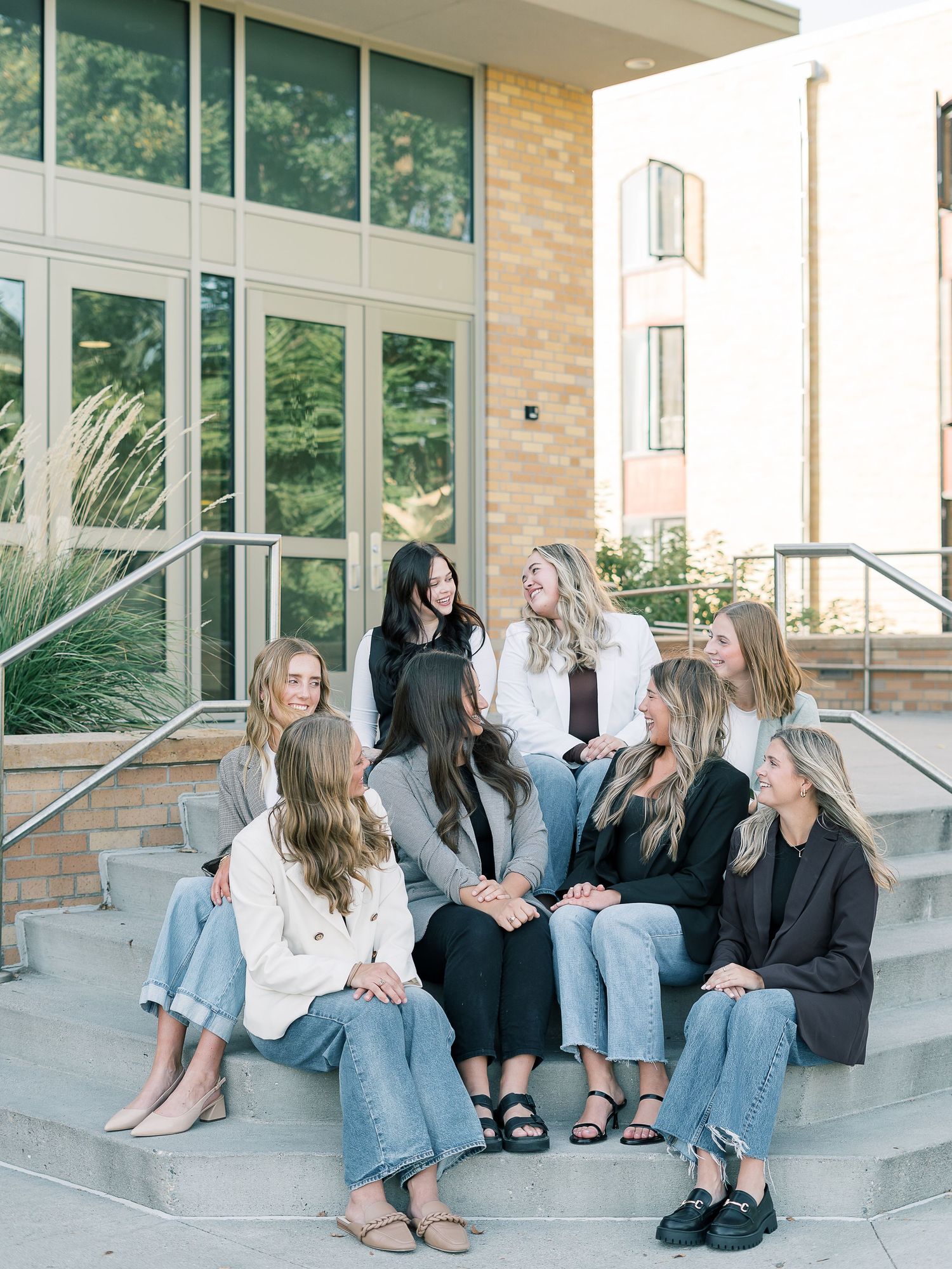 A group of friends sit casually together on concrete steps outside a modern building with glass windows and stone walls.