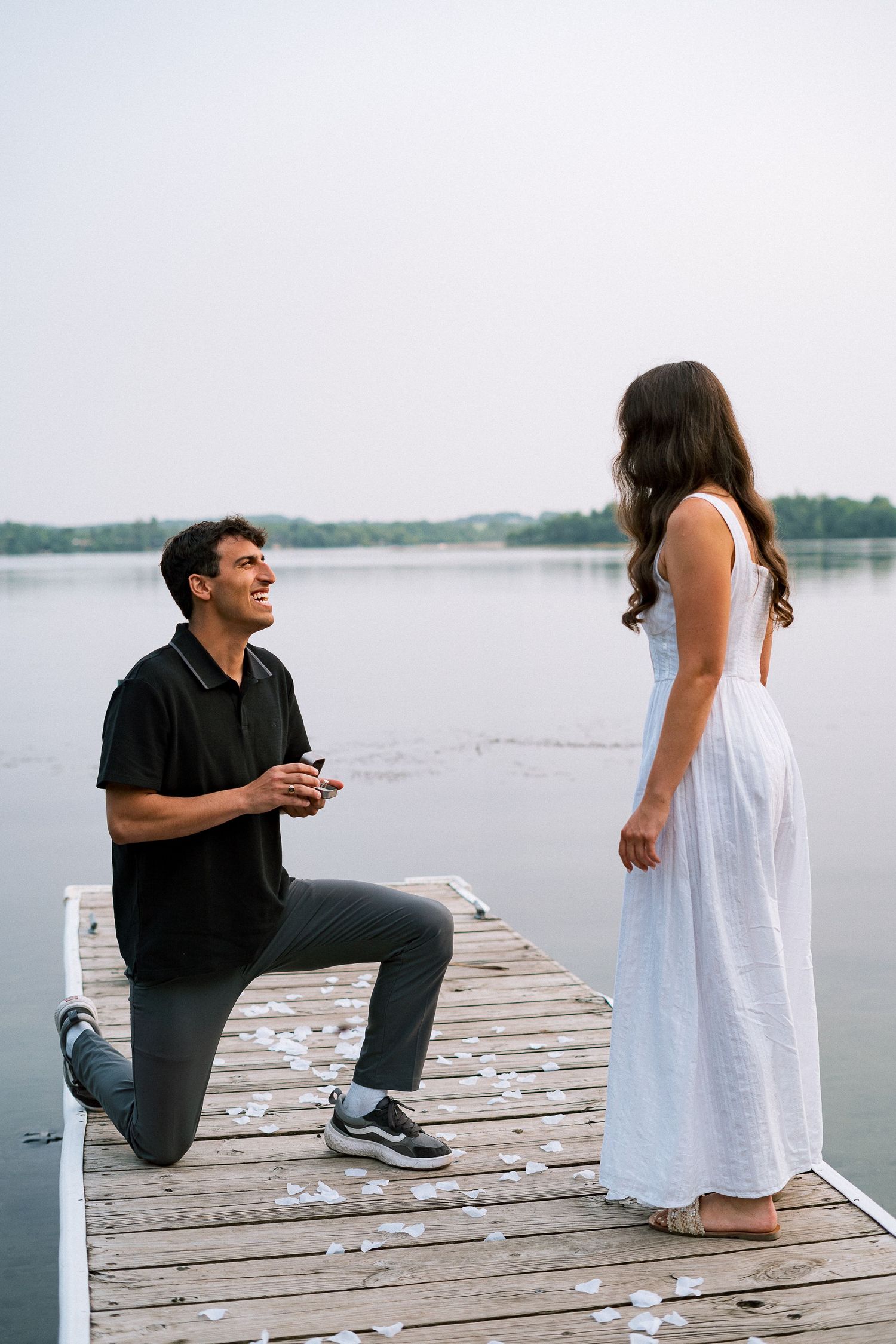 A romantic proposal scene unfolds on a wooden dock overlooking a serene lake.