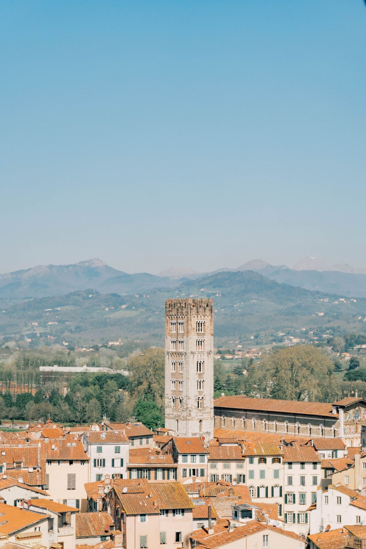 Panoramic cityscape view of Italian town with historic bell towers and terracotta rooftops against mountain backdrop.