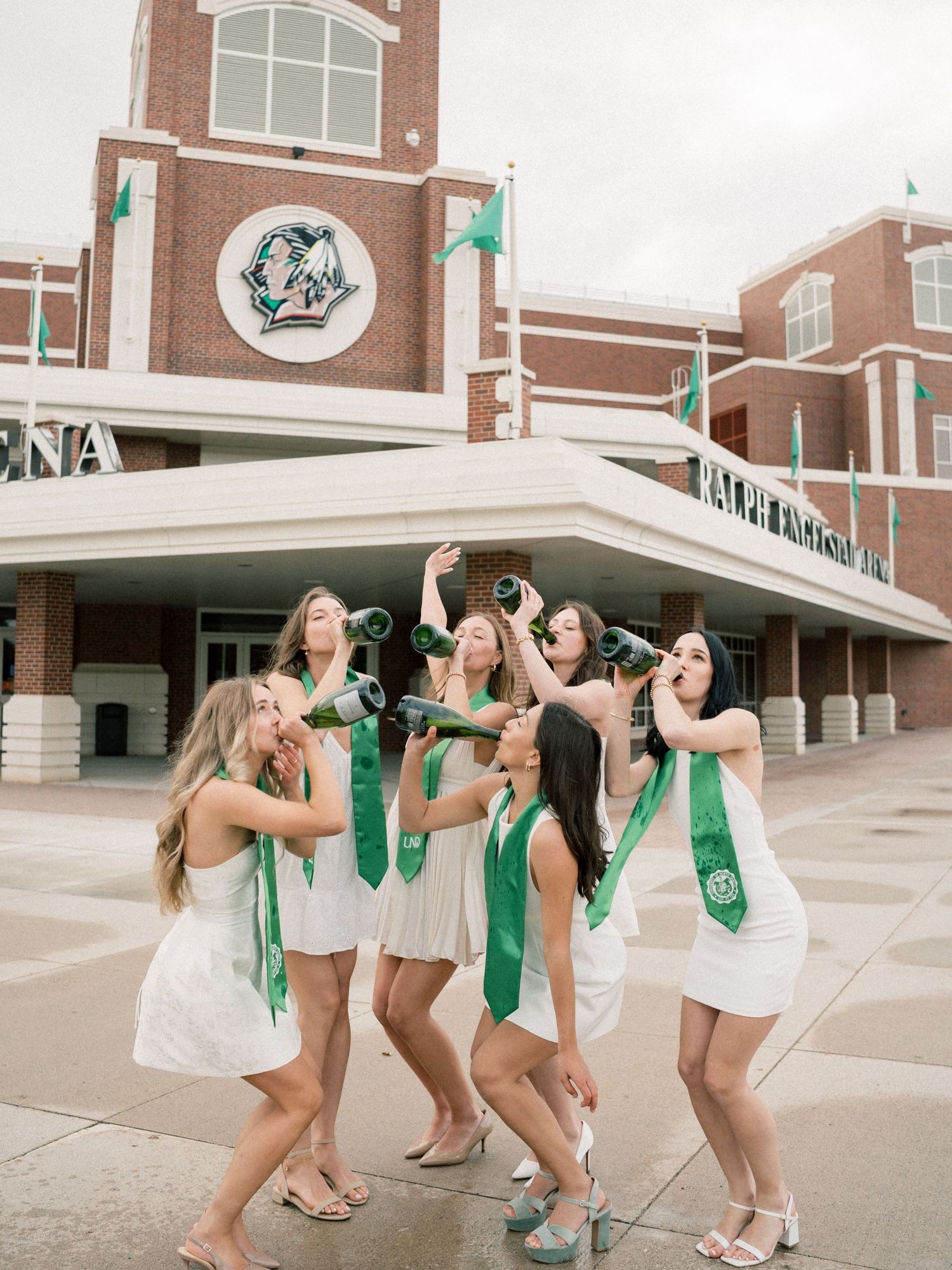 Group of college women in white dresses with green graduation stoles celebrating outside a brick university building.